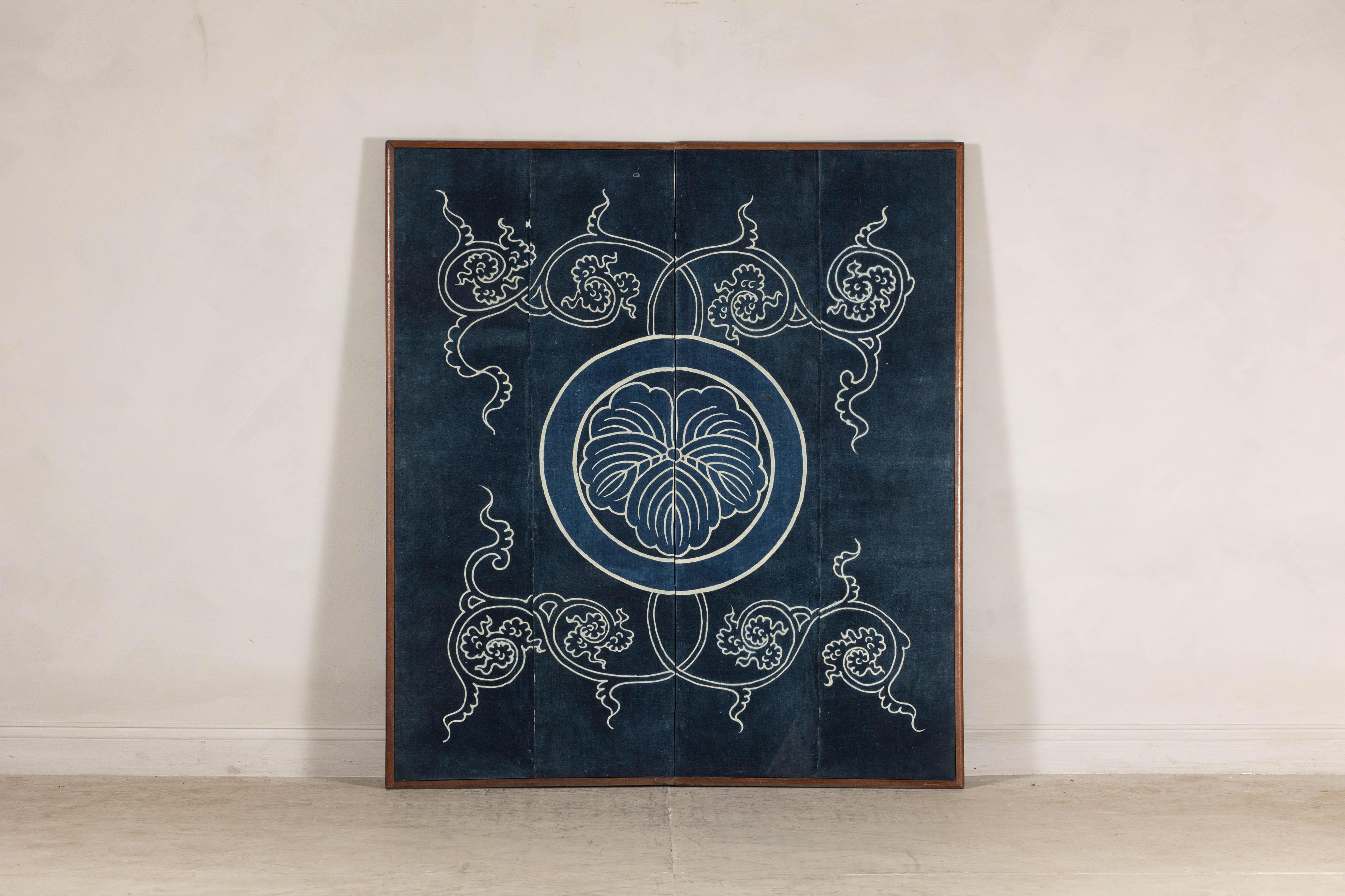 Un sorprendente pannello tessile indaco giapponese, incorniciato per l'esposizione, con un audace motivo a cresta reso in blu profondo e avorio morbido. Al centro, un medaglione circolare stilizzato evoca la formalità di un emblema mon o familiare,