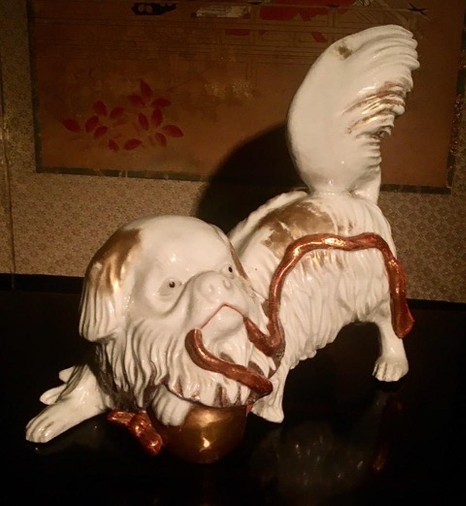 Japanese Kutani porcelain sculpture of a Pekinese dog, playing with a Shinto shrine bell and wearing a gold ruffle collar. The very expressive figure features a luminous white ground with gold accents and a bright bell rope. A rare and fine