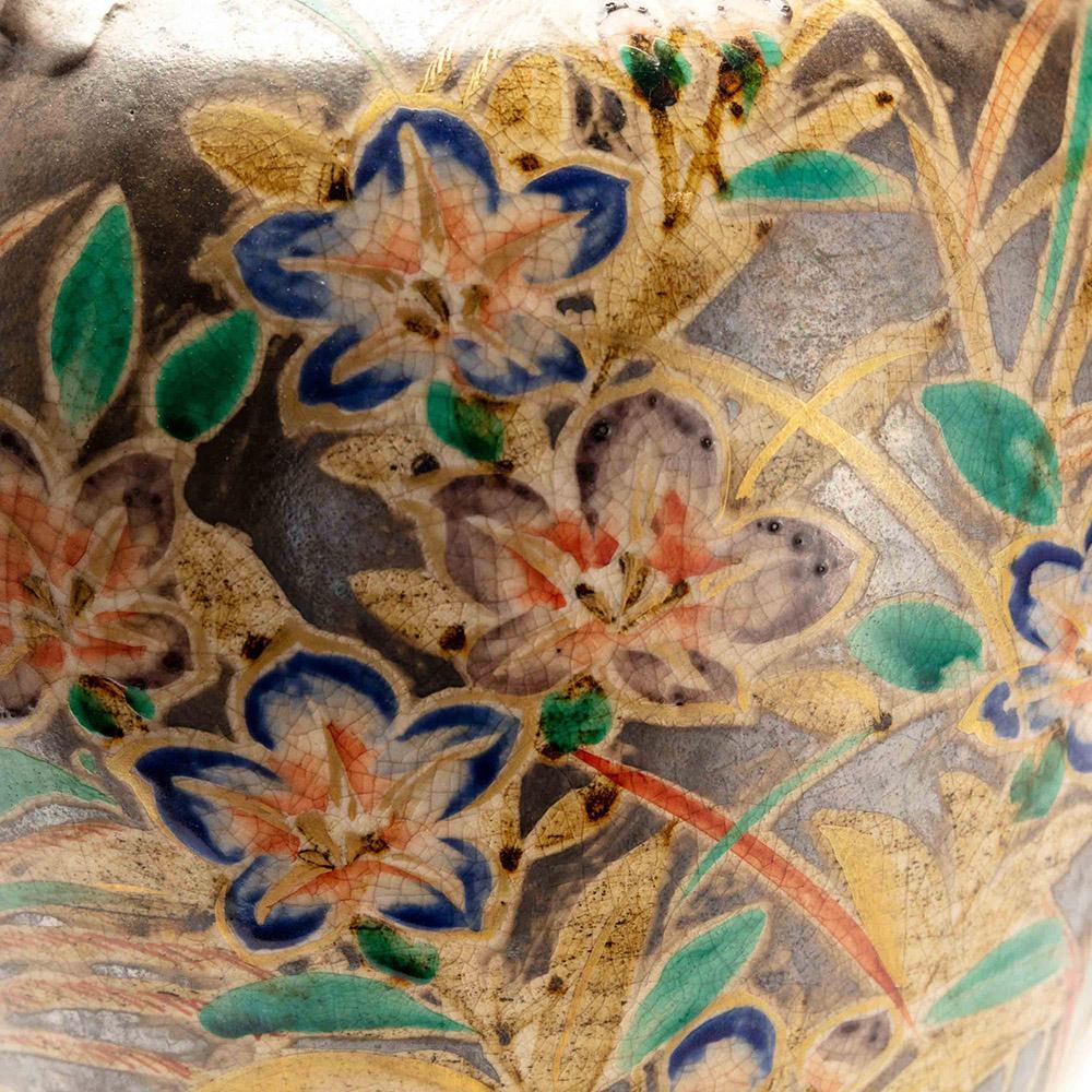 This Japanese Kenzan style vase has a classical shape with four small round handles. It has a colourful floral design of flowers and grasses. These have been hand painted in blue, green, purple and orange, on a silver gilt background that has a
