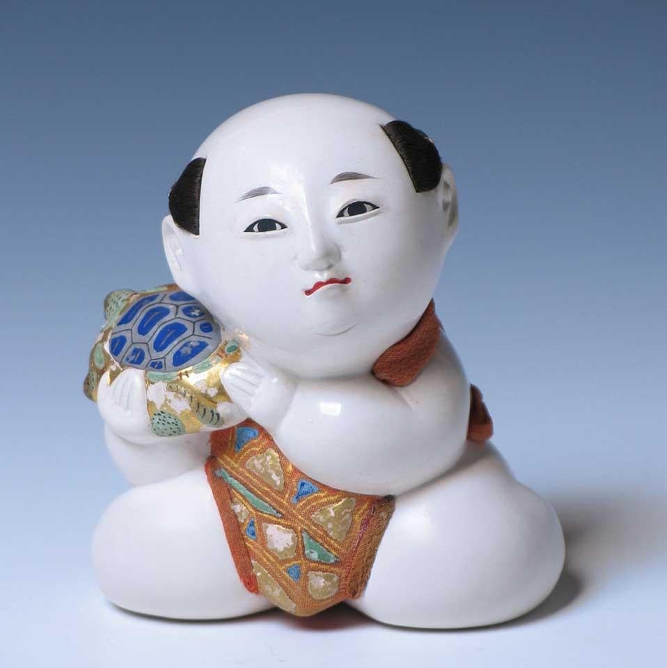 Japanese Kofuku-no-inori gosho ningyo (a good luck wish palace doll) of a plump kneeling male child holding a minogame, the longevity turtle, over his right shoulder, traditionally given amongst the nobility as a wish for a long life, all carved of