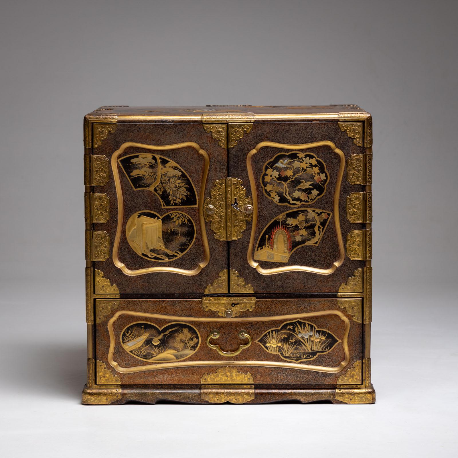 Small Japanese lacquer cabinet from the Meiji period with two doors and a drawer as well as side handles. The cabinet is decorated with gold lacquer decor (hiramakie) in the form of trees, landscapes, a carriage, a tent and small birds. The top