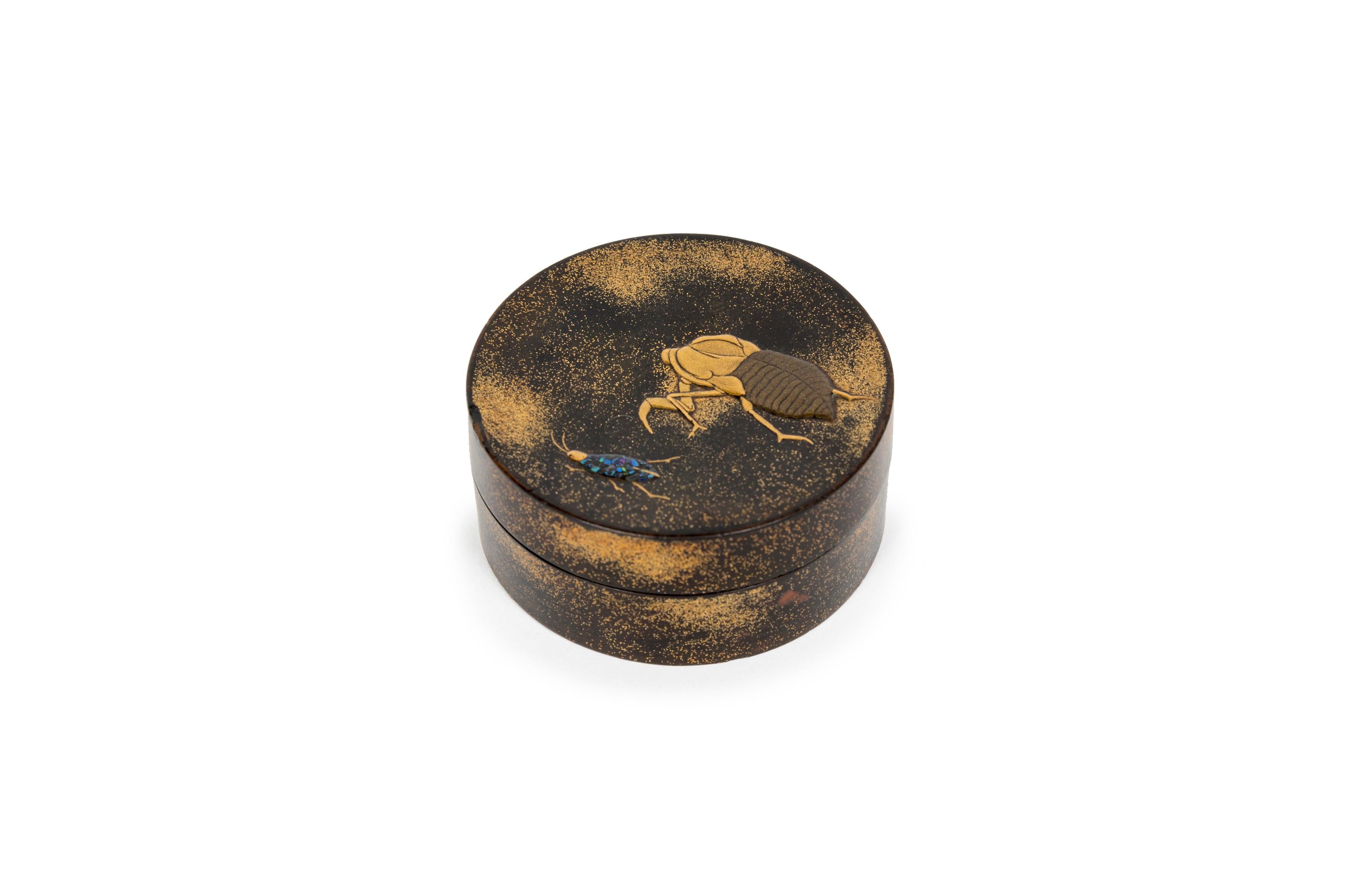Small Kobako circular box in lacquered wood with a cicada and unidentified insect, probably a gryllotalpa orientalis, displayed on its top part. 
The background is decorated in maki-e, creating a gold spray on black, and the cicada’s body is