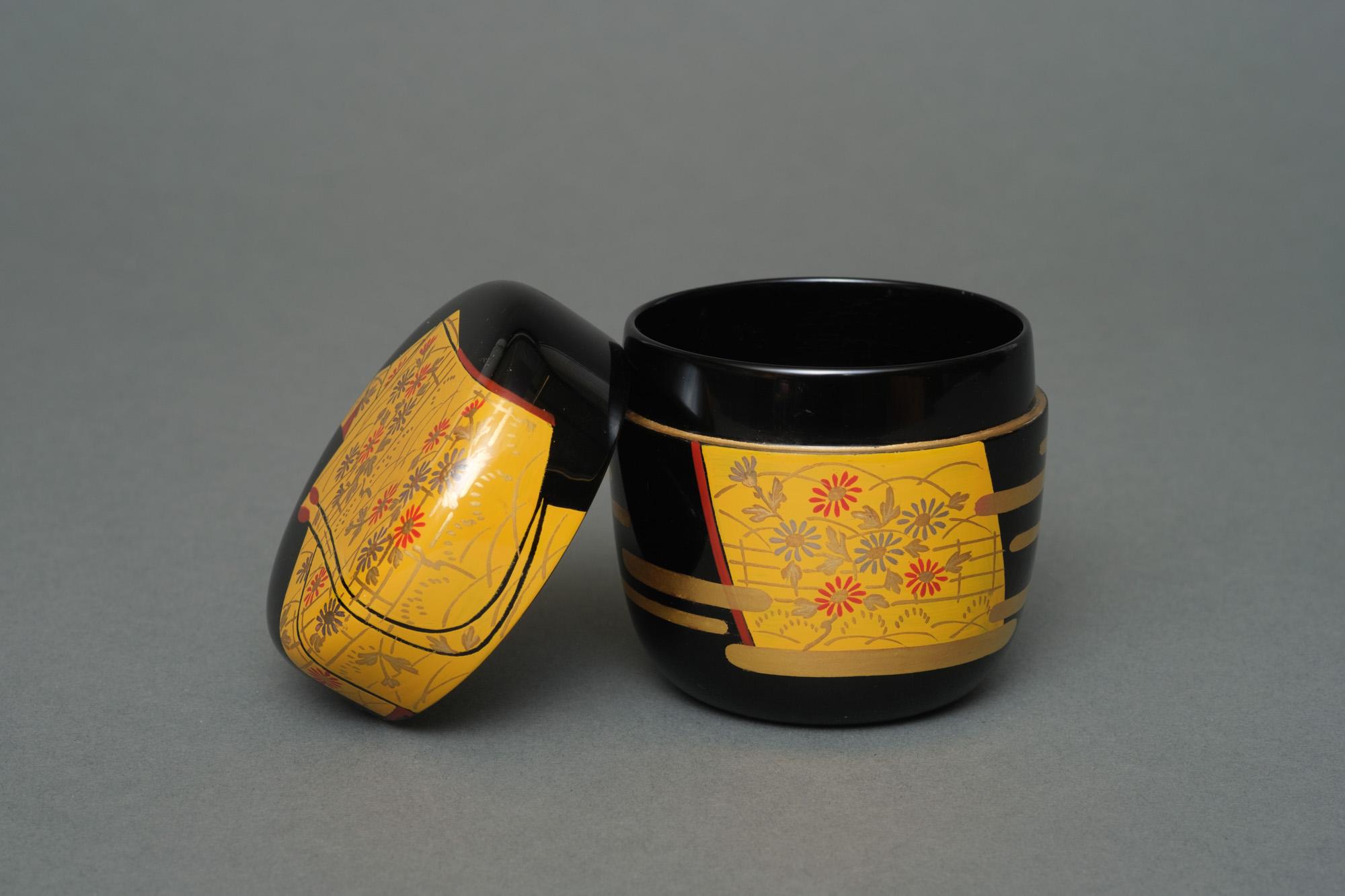 Hübsche Teedose (Natsume) aus schwarzem Lack mit einem farbenfrohen Hiramaki-e (Flachreliefmuster) eines gefalteten gelben Kimonos mit einem silbernen und roten Chrysanthemenblütenmuster. Der Sockel mit einigen goldenen stilisierten