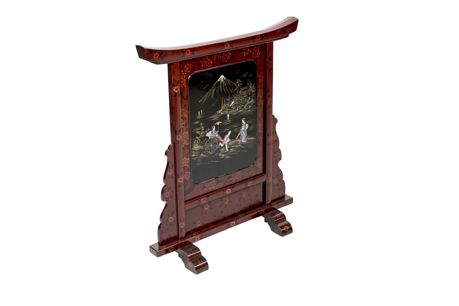 Japanese lacquered fire screen has a dark red lacquer background with floral borders around a central panel with painted mother of pearl figures. The figures depict man pulling a woman in a rickshaw with Mt. Fuji in the background. It is inlaid with