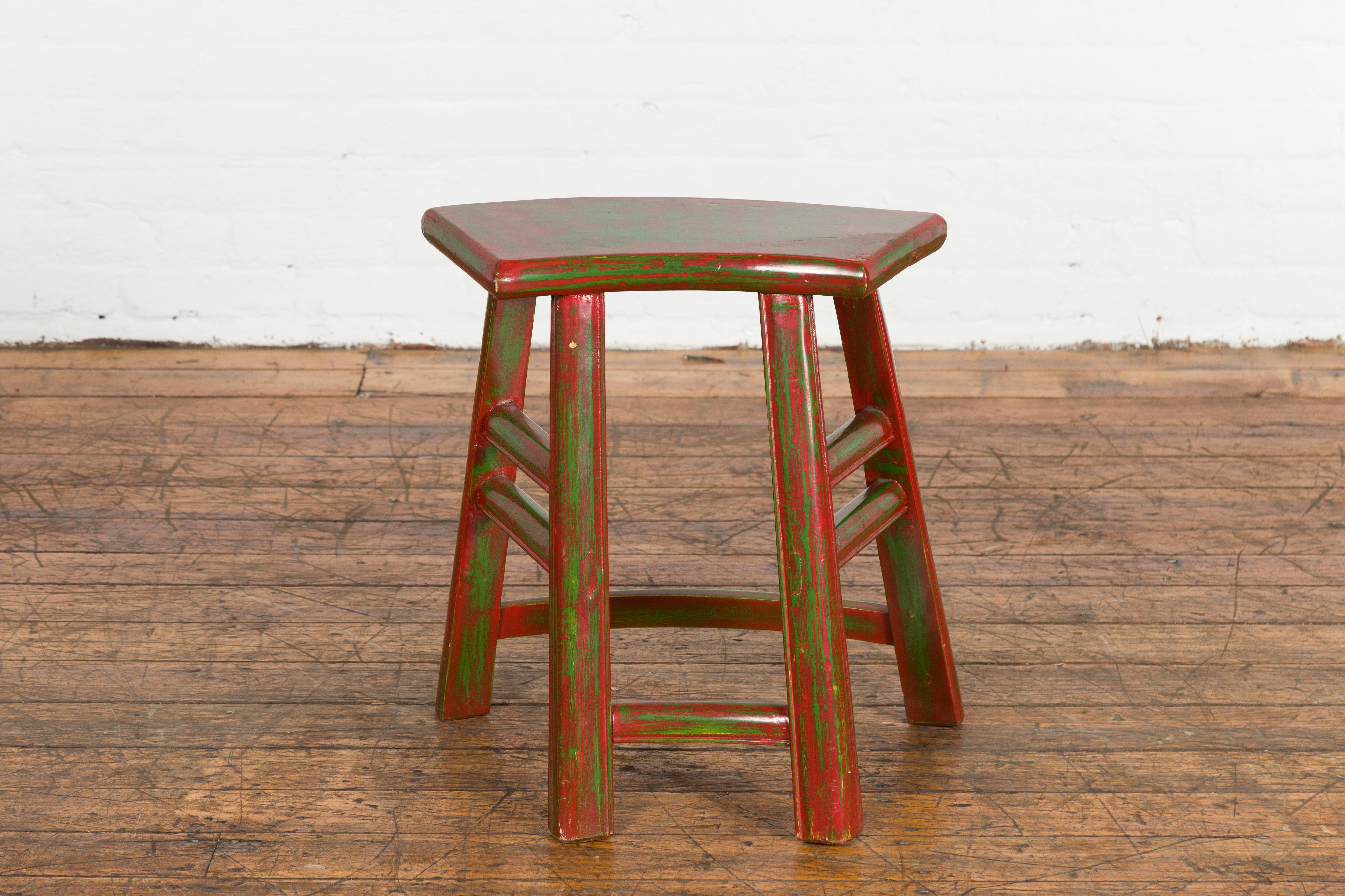 A Japanese late Meiji period red lacquered stool from the early 20th century, with hand painted green accents, semi circular top, four splaying legs and side stretchers. Created in Japan during the late Meiji period in the early years of the 20th