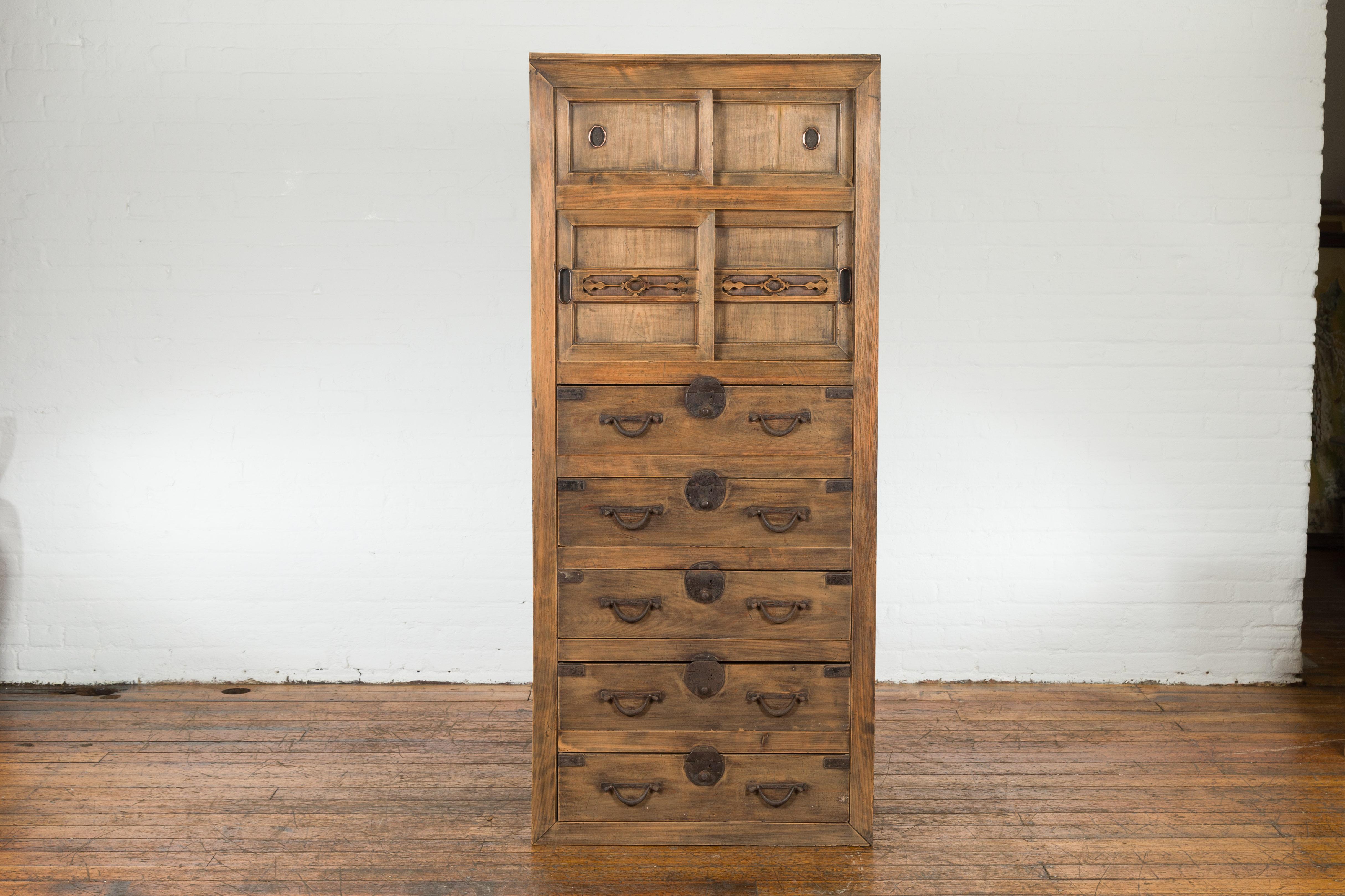 An Antique Japanese Meiji period Tansu clothing chest from the early 20th century, with iron hardware, carved panels, sliding doors, and five drawers. Created in Japan during the late Meiji period in the early years of the 20th century, this tall