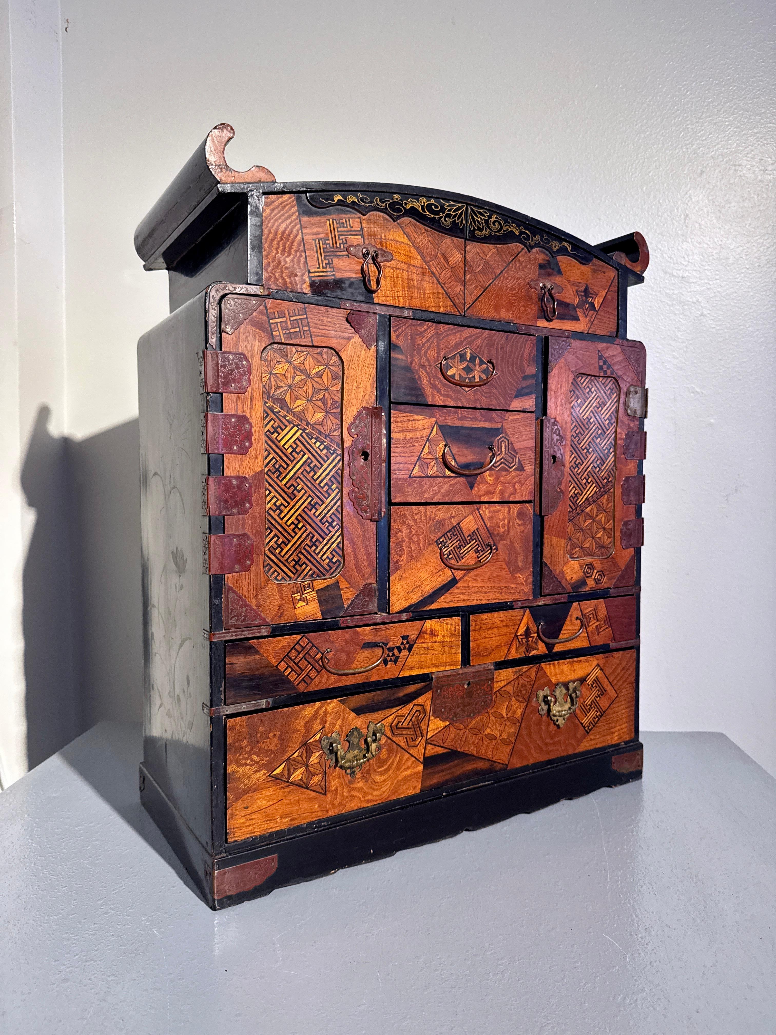 A large and remarkable Japanese lacquer and marquetry decorated table or hanging cabinet, Meiji Period, circa 1900, Japan.

The cabinet of square form surmounted by an arched top with upturned scroll ends. Eight drawers of various shapes and sizes