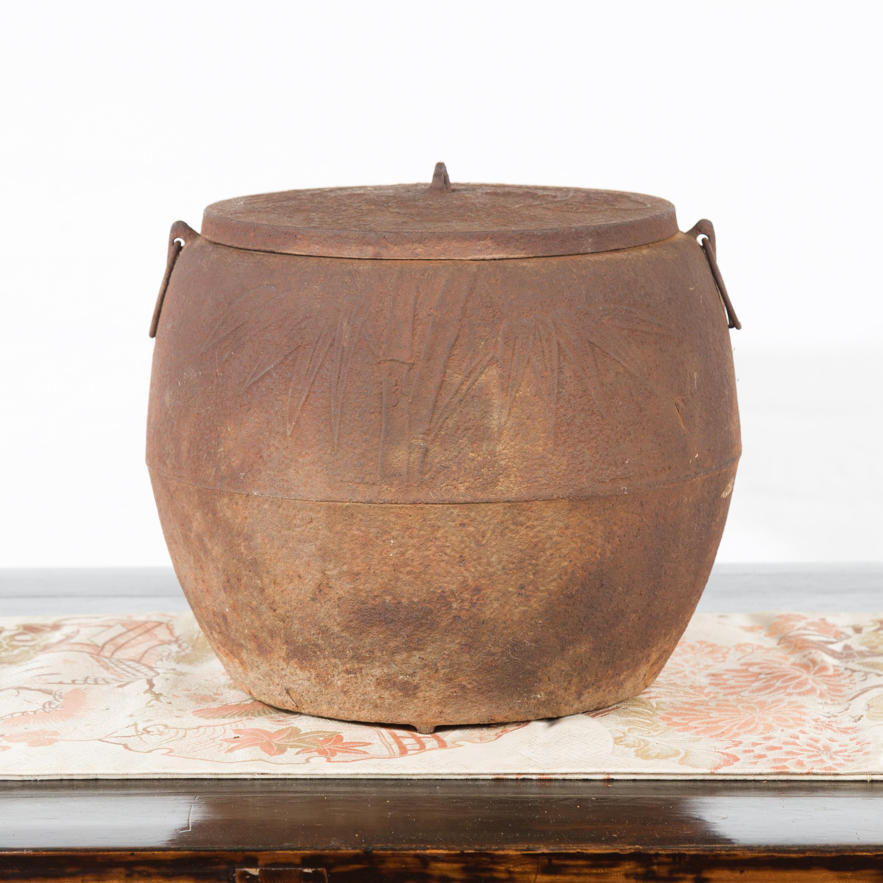 A Japanese Meiji period iron cooking pot from the early 20th century, with foliage motifs and distressed patina. Created in Japan during the early years of the 20th century, this iron cooking pot features a rusty circular body topped with a lid.