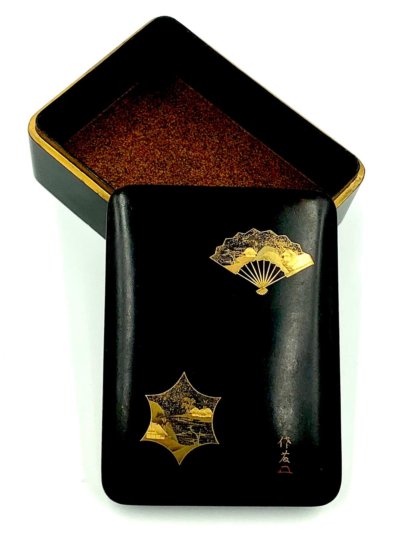 A fine antique Japanese Meji Period lacquer box decorated with a fan and a stylized star shaped window in the maki-e technique. The fan at the top right corner, fully opened depicting an idyllic traditional Japanese landscape with two charming