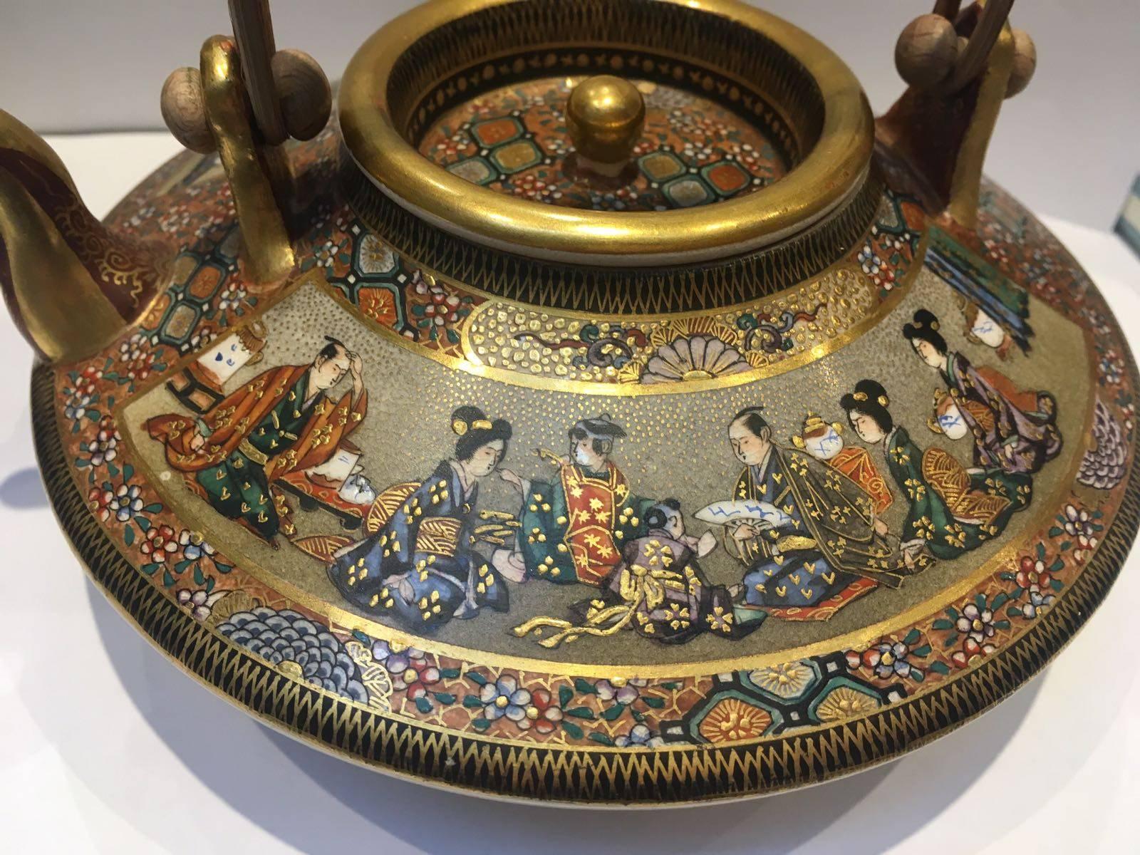 A Japanese Meiji Period Satsuma tripod sake pot with a bamboo handle. Intricately decorated, the pot features interiors scenes and various patterns filling in the porcelain flattened teapot shape. Gilt, the pot is further decorated with floral