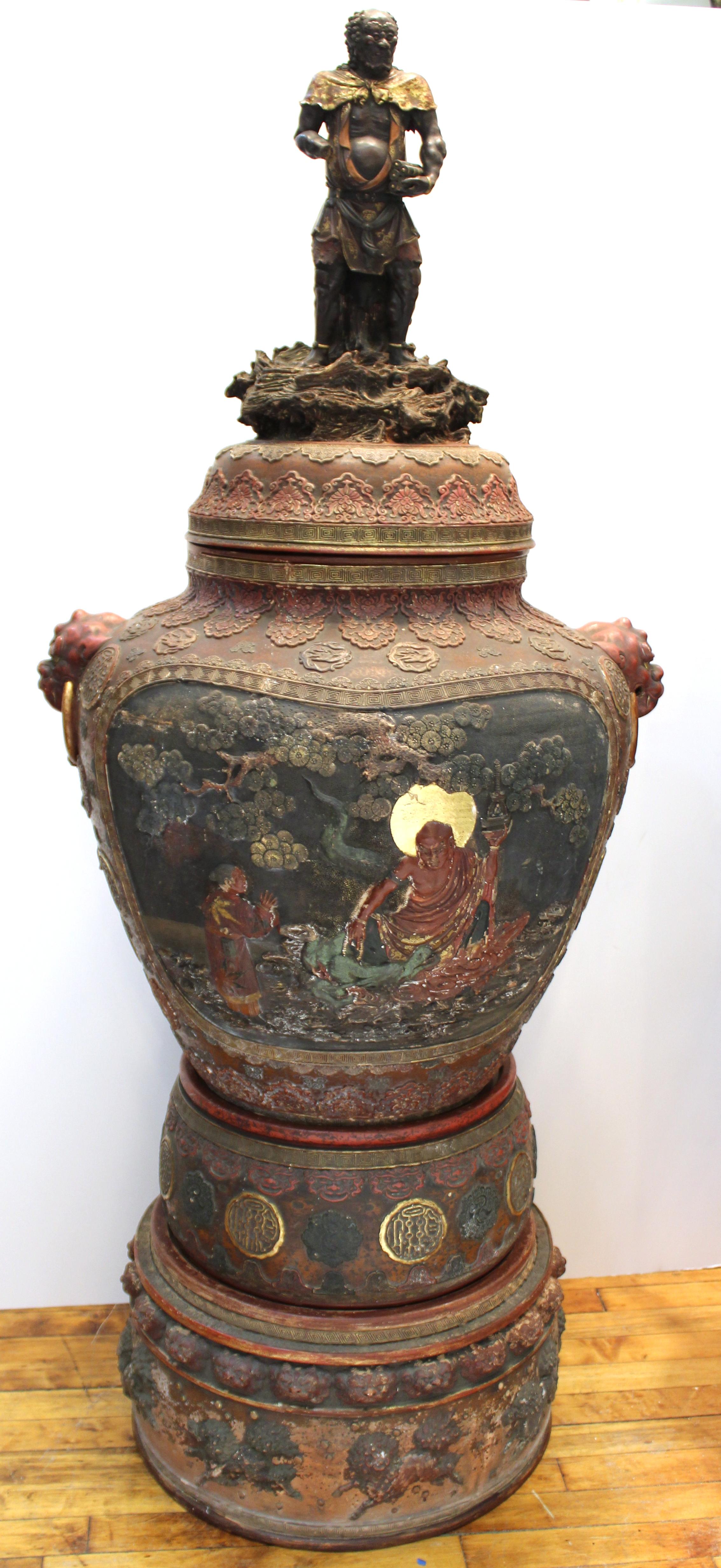 Japanese Meiji Satsuma monumental polychrome vase in terracotta with sculpted lid atop multiple layered Stand. The elaborately decorated piece features large medallions on the front and back with scholarly scenes and grotesque masks as handles on