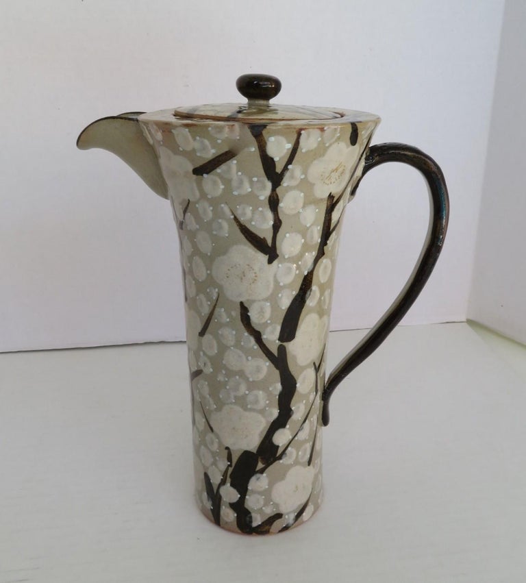 Exquisite Mid-Century Modern Japanese tall and slender ceramic pitcher with top.  Decorated with stylized cherry blossom branches, with white flowers on a gray green background. Lovely form.  Very Good condition with minor wear and no issues. 