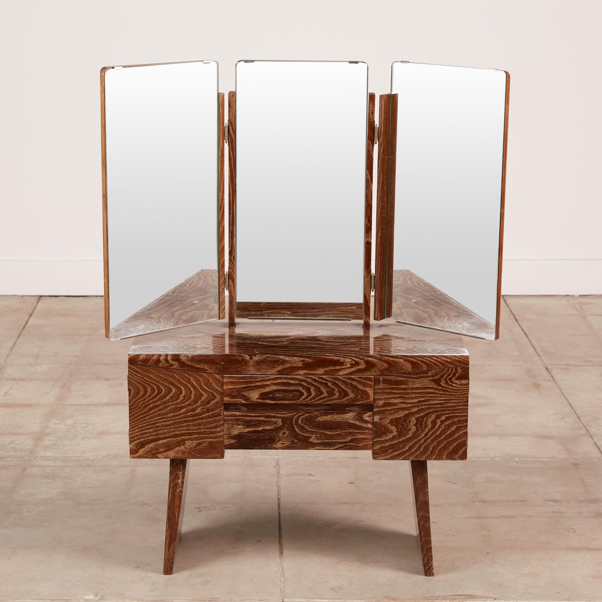 A petite Japanese modernist vanity with tri-folding mirrored panels that sit atop a case with opposing doors and a middle pair of drawers. The cabinet is supported by a pair of tapered legs. The exterior is a stained wood grain with a glossy lacquer