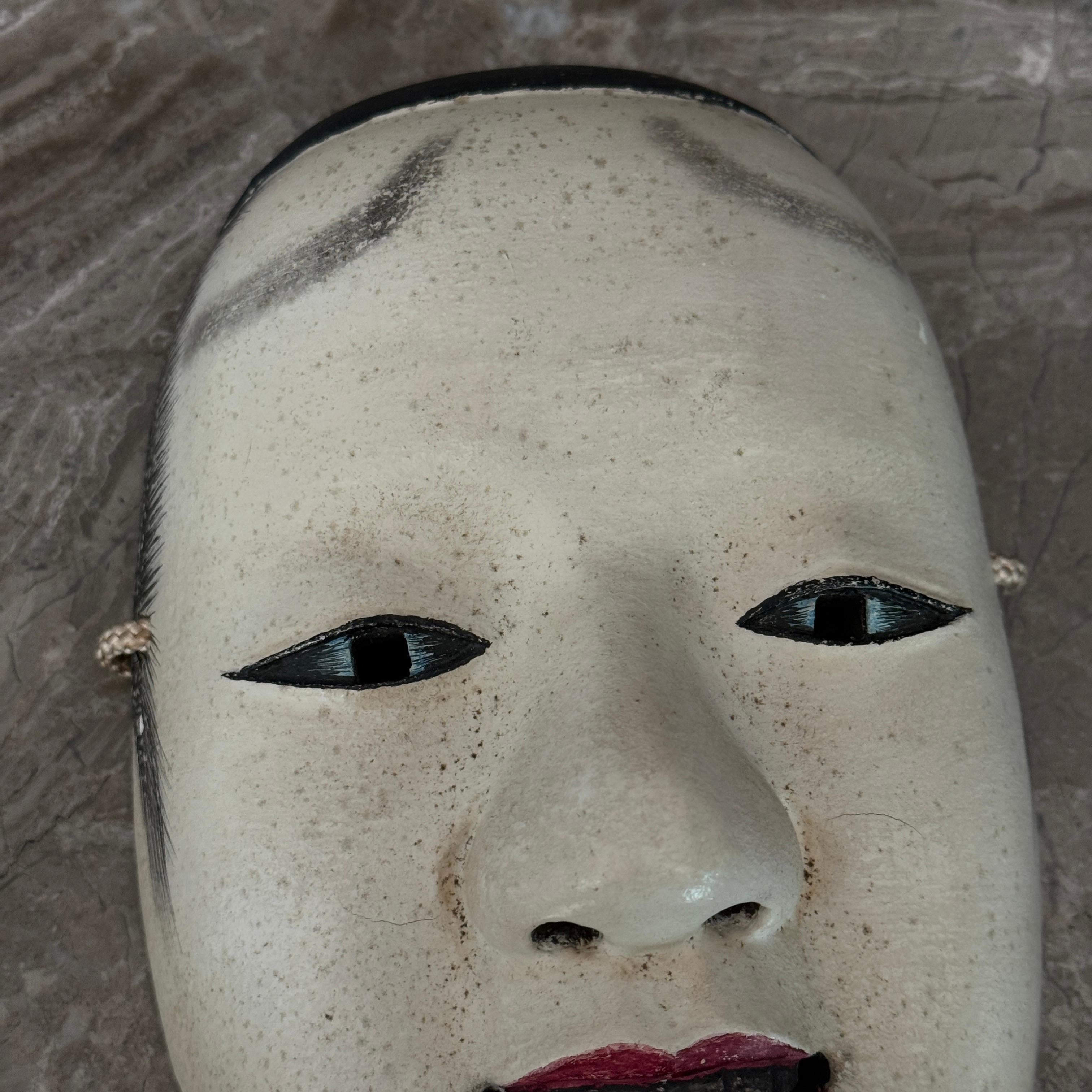 Ce masque japonais de nô est un exemple d'art théâtral traditionnel finement travaillé, sculpté à la main dans du bois et présentant une expression sereine et énigmatique typique des représentations classiques de nô. Le masque présente des détails