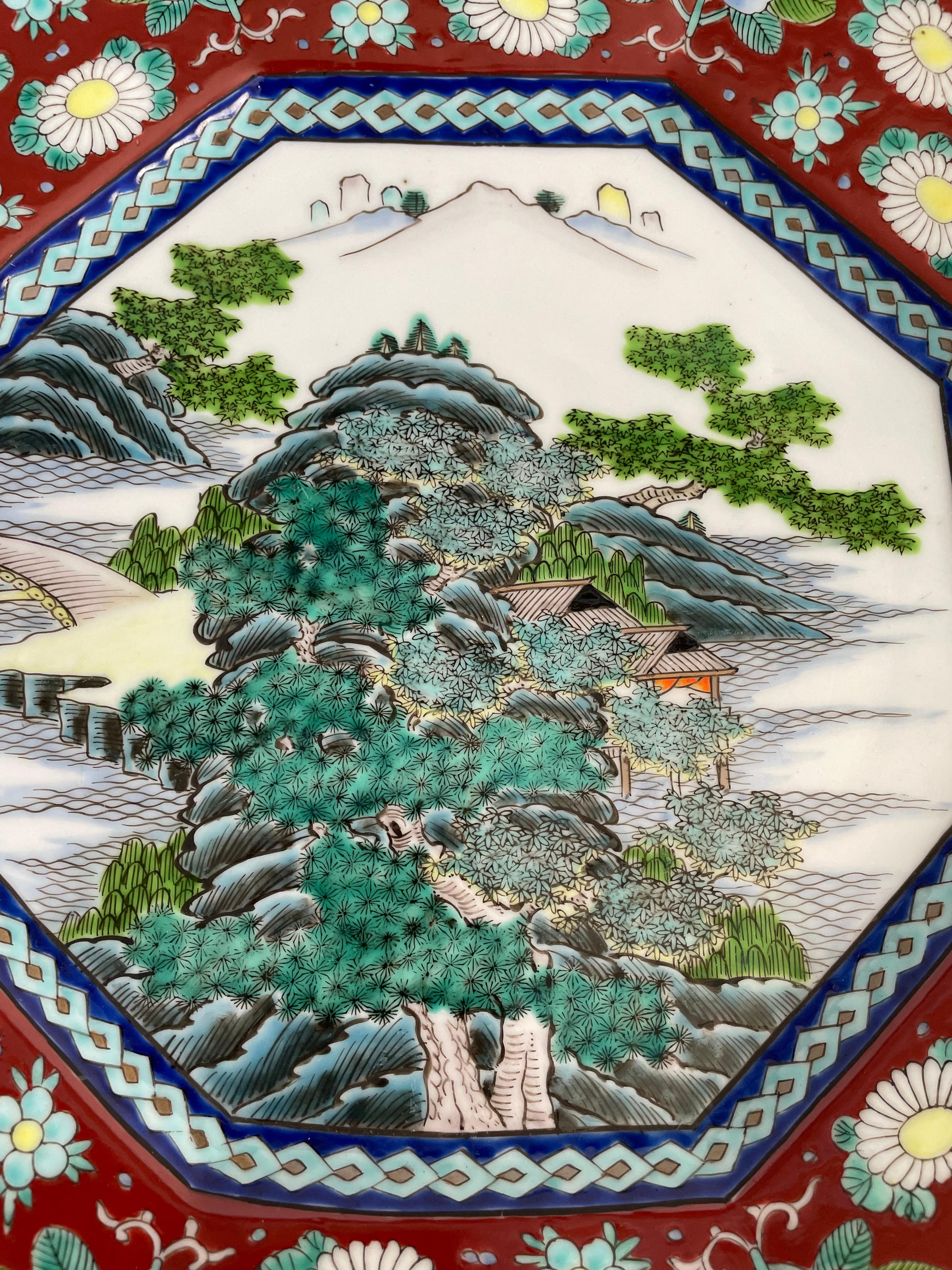 A decorative handpainted Japanese Imari octagonal ceramic charger, gilt edges, profusely decorated in vibrant shades or blue, red, green and yellow, featuring a rocky island with mature pine trees and a small shrine, connected by a causeway with