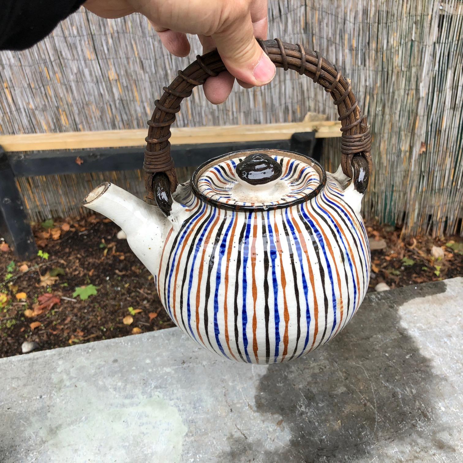 An attractive Japanese tea pot glazed in brilliant multi colored stripes in beautiful classic glazes: blue, black, and reds over white ground. The sturdy rattan handle completes this two-piece beautiful work of art.

Good condition and ready to