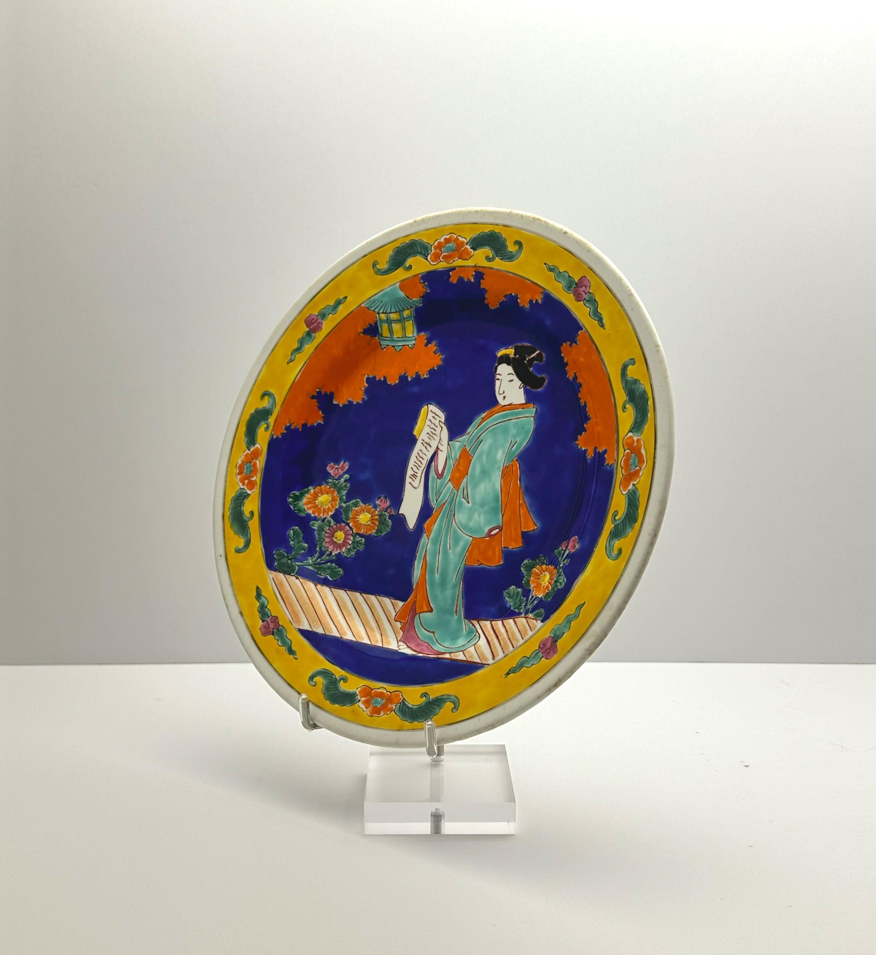 A bold blue and yellow decorative porcelain charger plate, hand-crafted in Japan around the 1920s.

Hand-painted with a scene depicting a Geisha reading from a scroll on a walkway adorned with chrysanthemum flowers, this decorative plate features a