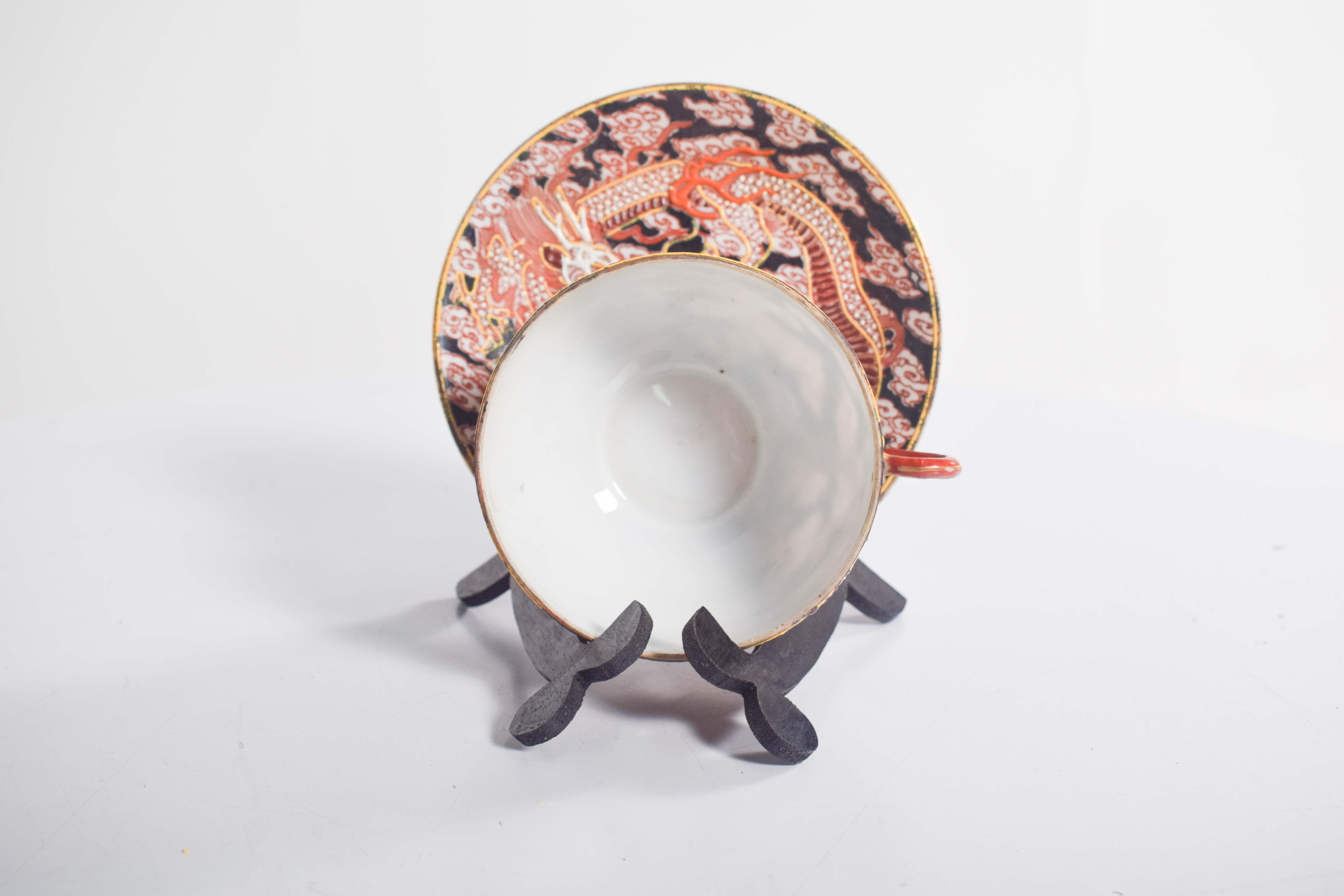 This exquisite Japanese porcelain cup and saucer set showcases vibrant polychrome decoration, blending rich red, black, and gold tones. The centerpiece of the design features a coiled snake or dragon motif, surrounded by swirling clouds and golden