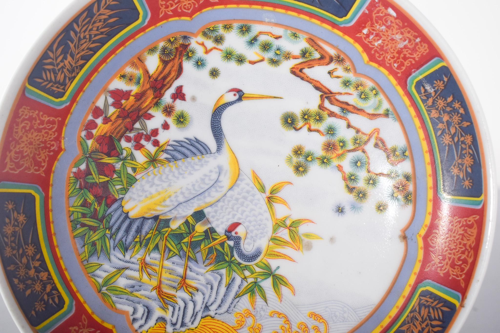 Assiette en porcelaine japonaise. Exubérante décoration polychrome richement ornée de couleurs et d'éléments jaunes, rappelant l'or. Au centre se trouve une illustration détaillée de deux élégantes grues, l'un des symboles les plus traditionnels de