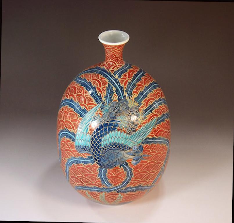 Stunning contemporary decorative porcelain vase showcasing a blue and turquoise Phoenix gracefully extending its wings all around the body of the vase on a red background with a detailed flower pattern in gold. Phoenix has since ancient times been
