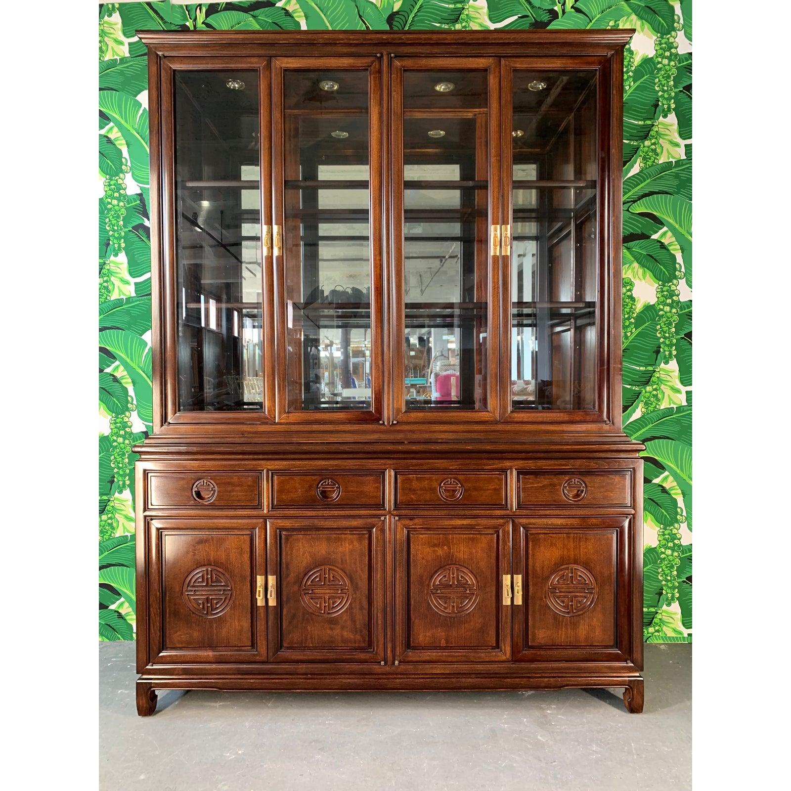 Asian chinoiserie display cabinet features solid redwood construction and brass hardware. Lighted interior with mirrored back with glass shelves. Hand carved drawer pulls and door faces. Very good vintage condition overall.