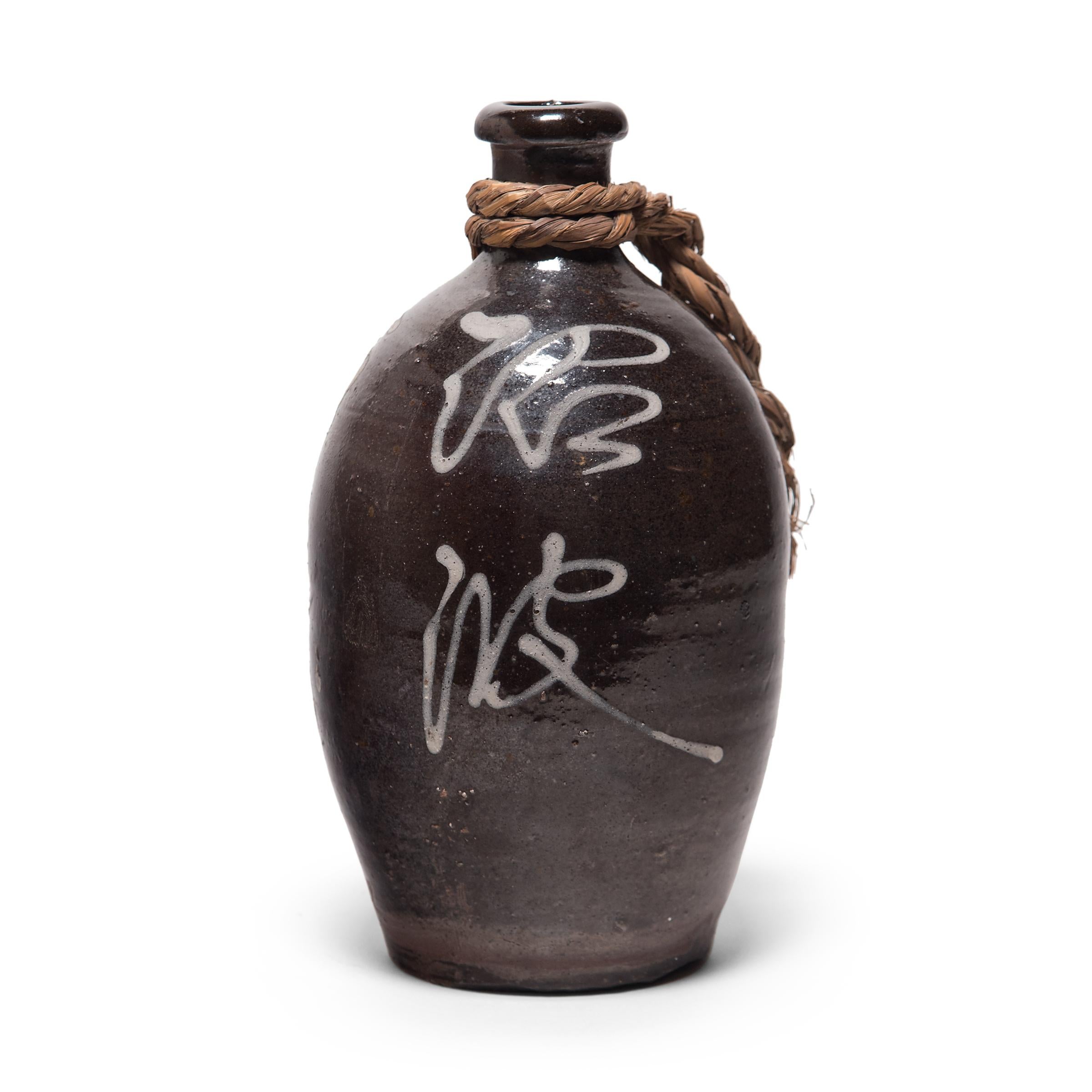 This glazed ceramic jar is a Japanese sake bottle from the late-Meiji era. The jar has a rounded bottleneck form and is coated with a dark brown glaze in resemblance to bizen ware pottery. Applied in a contrasting light glaze, expressive calligraphy