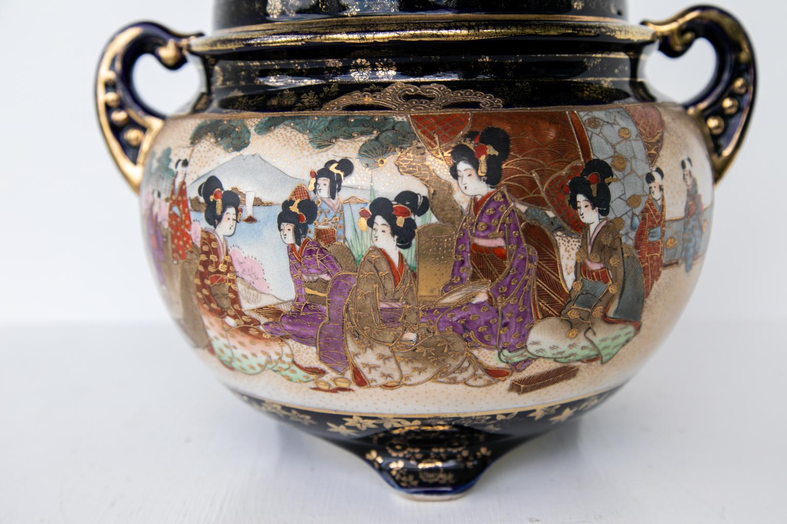 Japanese satsuma covered tureen, depicting figures in a garden and a courtyard. The background is cobalt blue with gilt highlights all-over. The lid has heart shaped cutout vents. There is some wear to the gilding.
        
