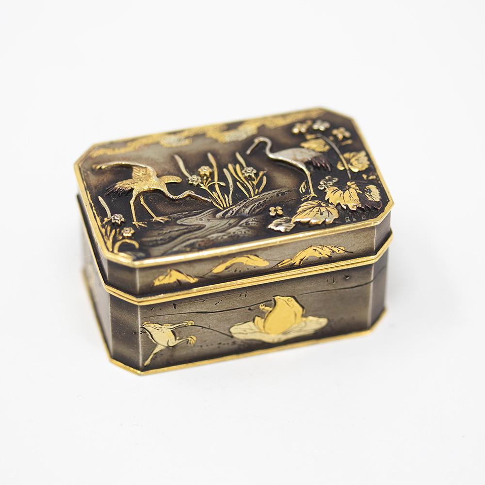 Fine Japanese Meiji period shakudo pill box decorated with wildlife of komai style damascene form. The box beautifully decorated with cranes to the lid above a flowing river beside blossoming flowers and foliage. To each longer side frogs jump