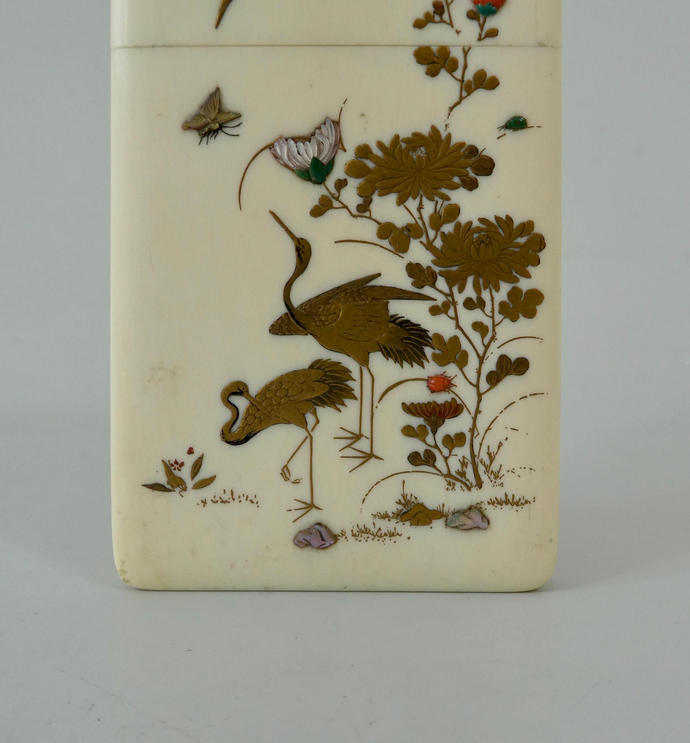 A Japanese Shibayama style card case, c. 1890, Meiji Period. The front of the case painted with cranes amongst flowering plants, and insects, decorated in gilt and inlaid hardstones. 
The reverse painted in gilt lacquer, with two panels, with scenes