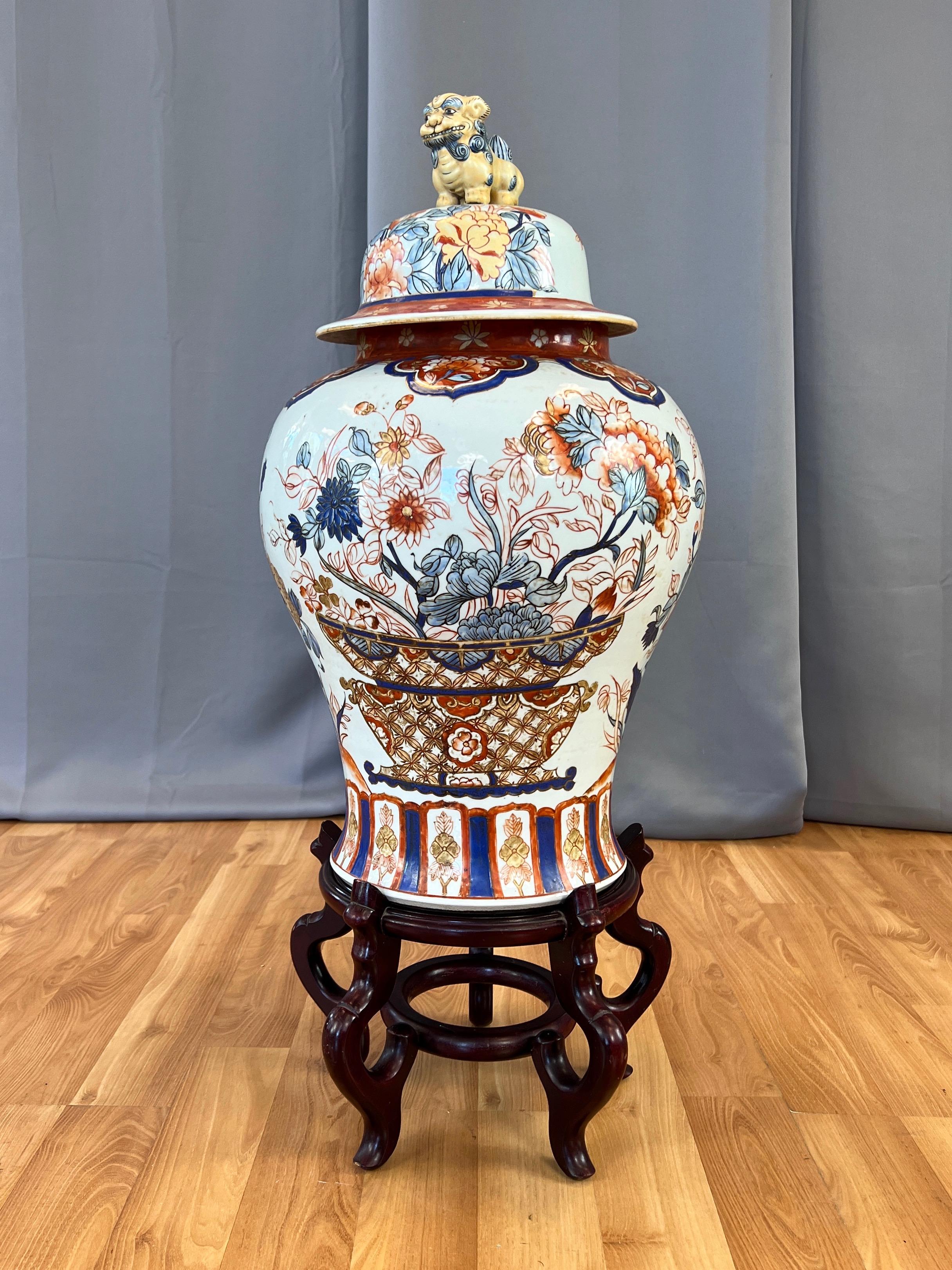 A circa 1940 extra-large Japanese Shōwa era Imari ware hand-decorated porcelain lidded temple jar, urn, or vessel on wood stand.

Lid features a delightfully charming butter yellow and baby blue grinning shisa figure on top—a traditional Ryukyuan