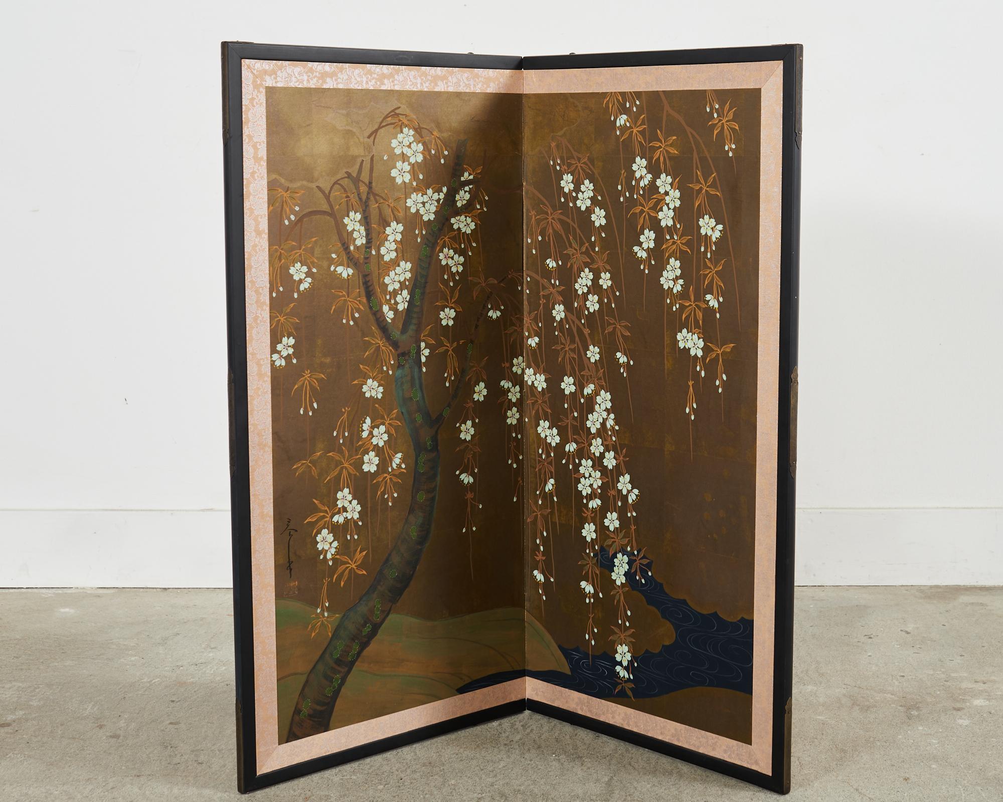 Gorgeous Japanese 20th century Showa period two panel byobu screen depicting a delicate flowering cherry (sakura) tree by a meandering river or stream. Painted with ink and natural color pigments over a gilt square ground. The delicate white