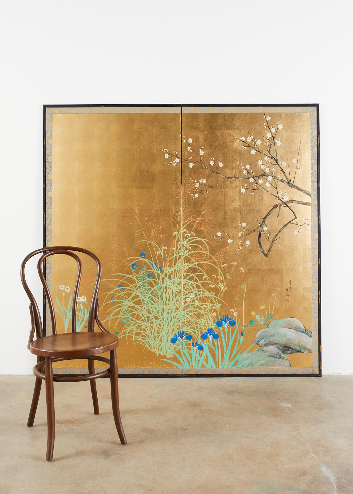 Large Japanese two-panel screen featuring a flowering landscape over a dramatic gold leaf background. The idyllic scene has a flowering plum tree above dandelion, iris, bellflower, and pampas grass. Ink and colored pigment over squares of gilt