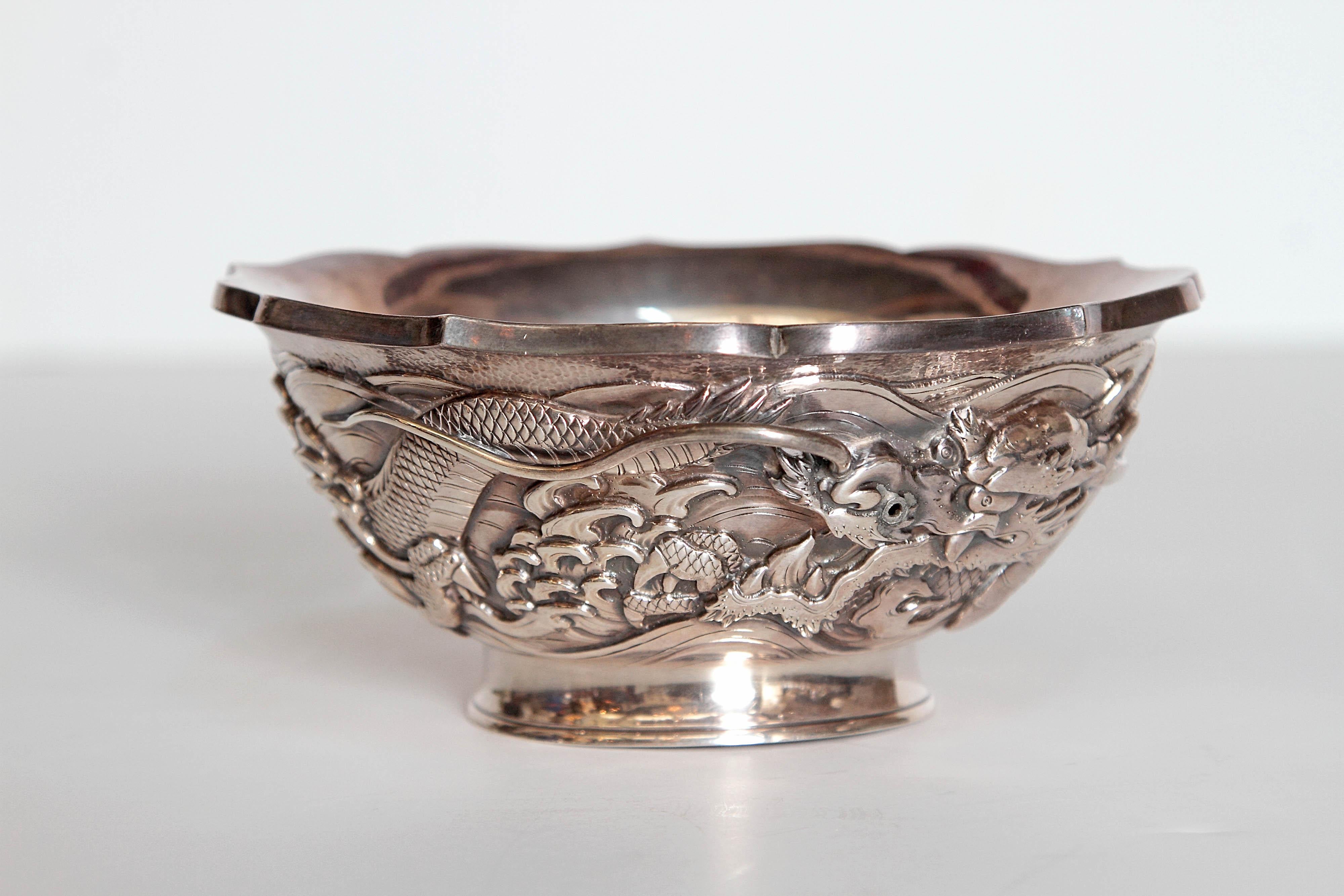 A Japanese silver bowl marked with highly decorative curving dragon in high relief around the body. Scalloped edge on top in circular form, side embossed with a dragon and scroll decoration, on a circular foot.