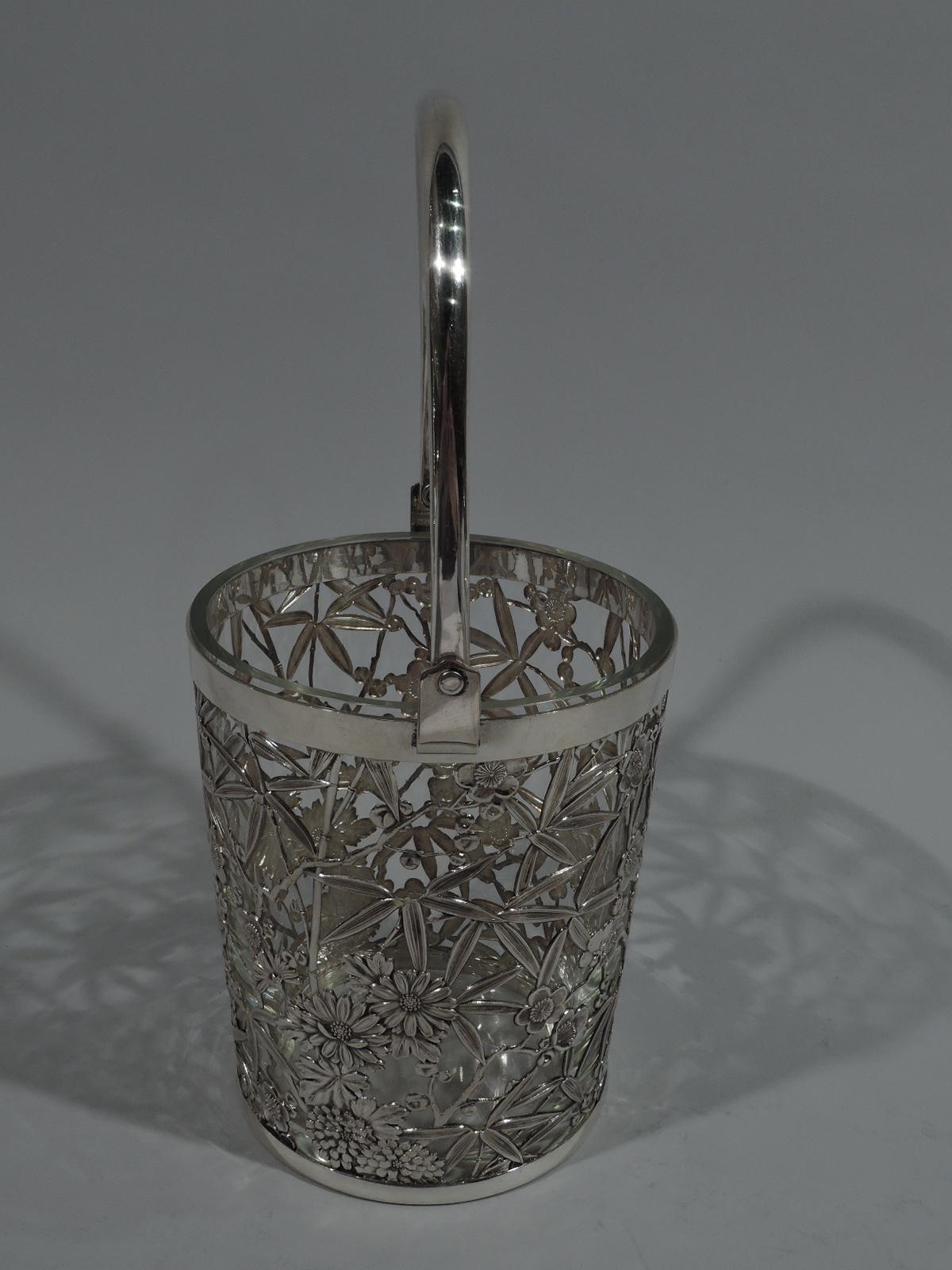 Japanese silver ice bucket. Round with straight sides and plain rim and vase. Loose floral openwork on sides with pell-mell blossoms interspersed with bamboo leaves. C-scroll swing handle. Detachable clear glass liner. Marked “Sterling 950”.