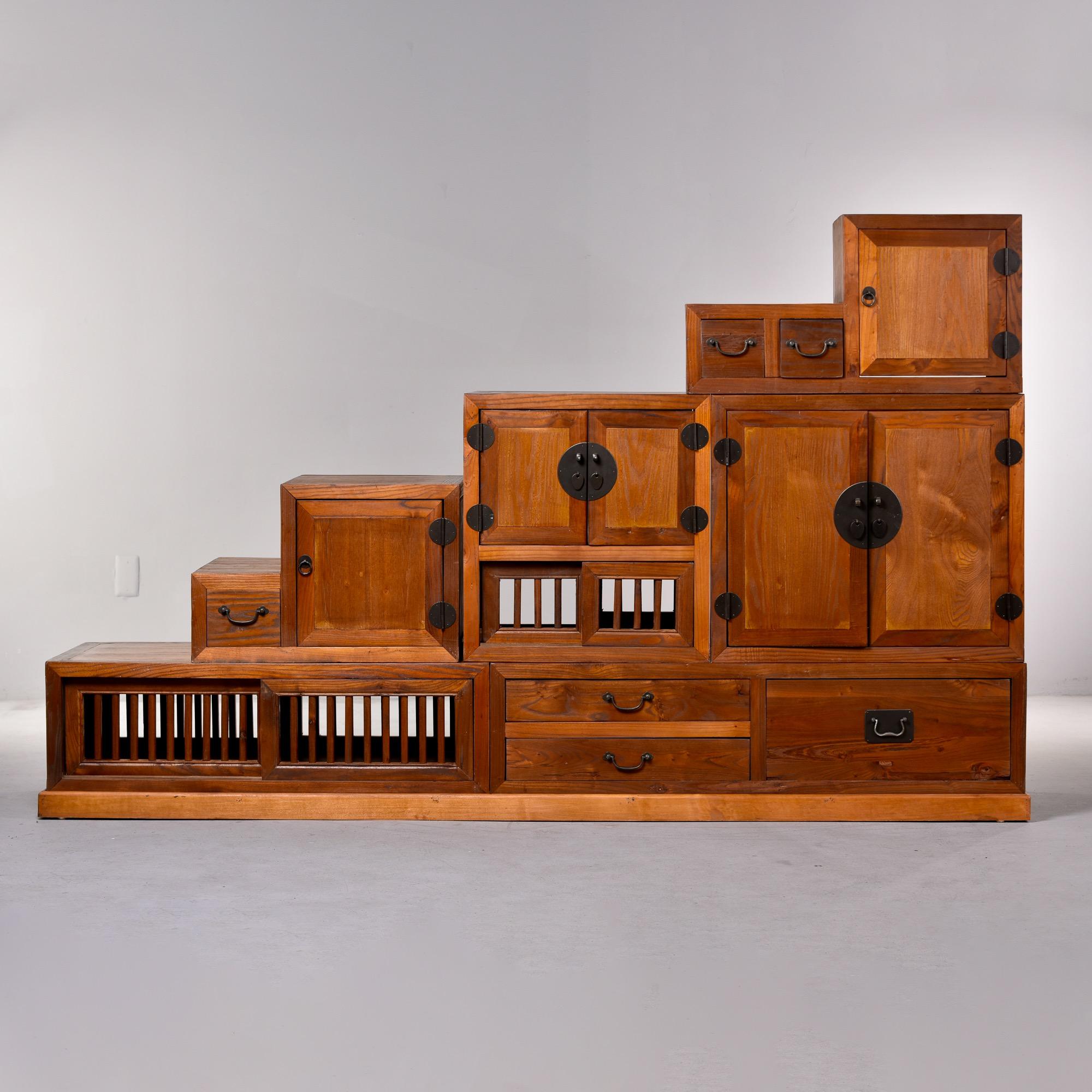 Circa 2020 Japanese two-sided Tansu style cabinets and base pieces in stained wood with black iron hardware. Components include ladder-like base and six cabinets of various sizes that can be arranged and stacked on the base in many configurations.