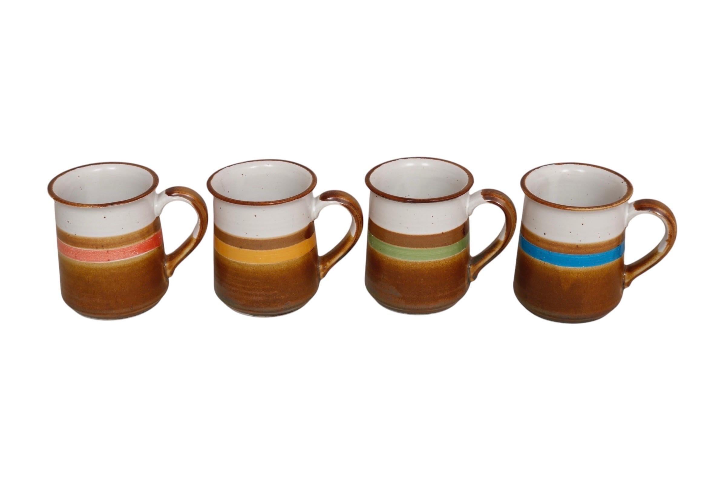 A set of four colorful stoneware mugs. Each is glazed with a speckled white and gloss brown glaze, separated with a band of color; coral, mustard, olive green and cerulean blue. Marked underneath ’Japan’. Dimensions per mug.