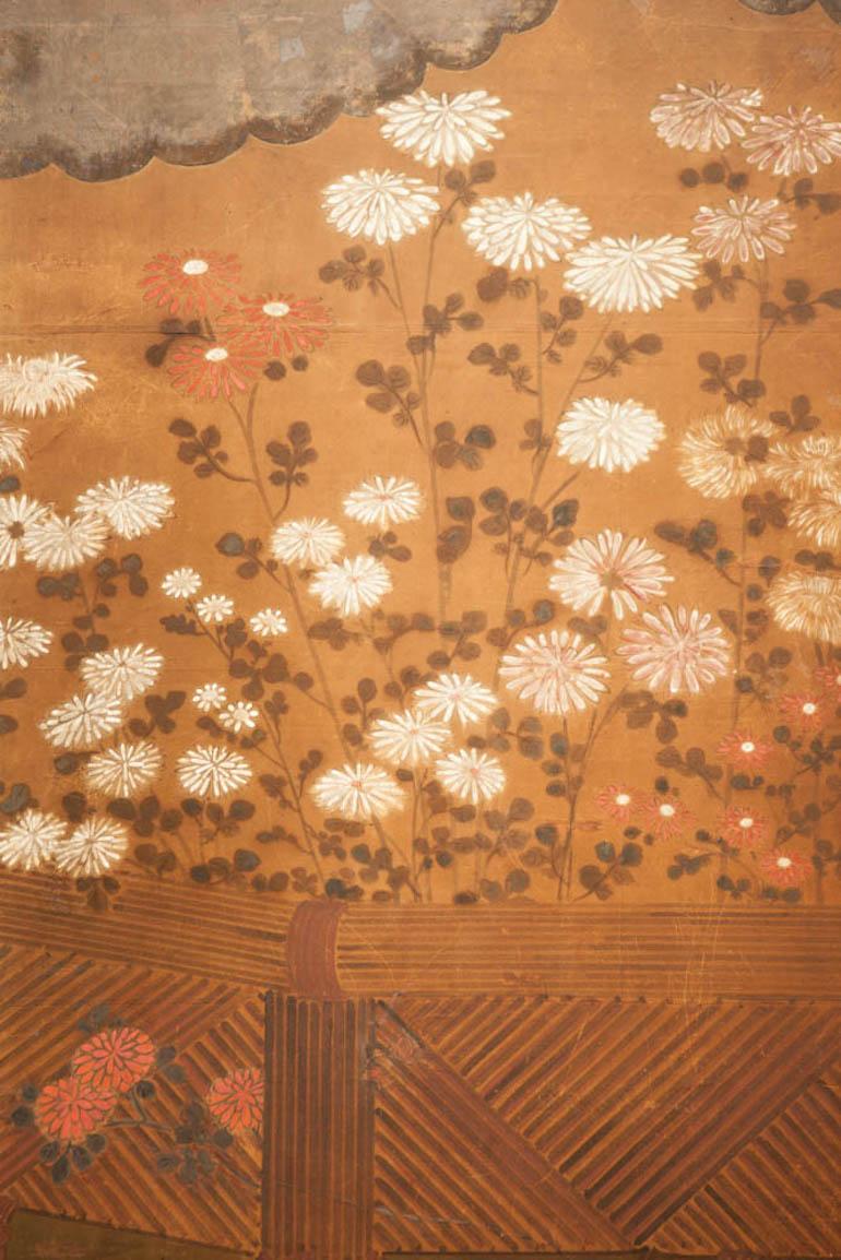 Japanese two-panel screen: Chrysanthemums over a woven reed fence, chrysanthemums growing behind and through a woven fence underneath silver leaf clouds. Mineral pigments on mulberry paper with oxidized silver leaf and a silk brocade border.