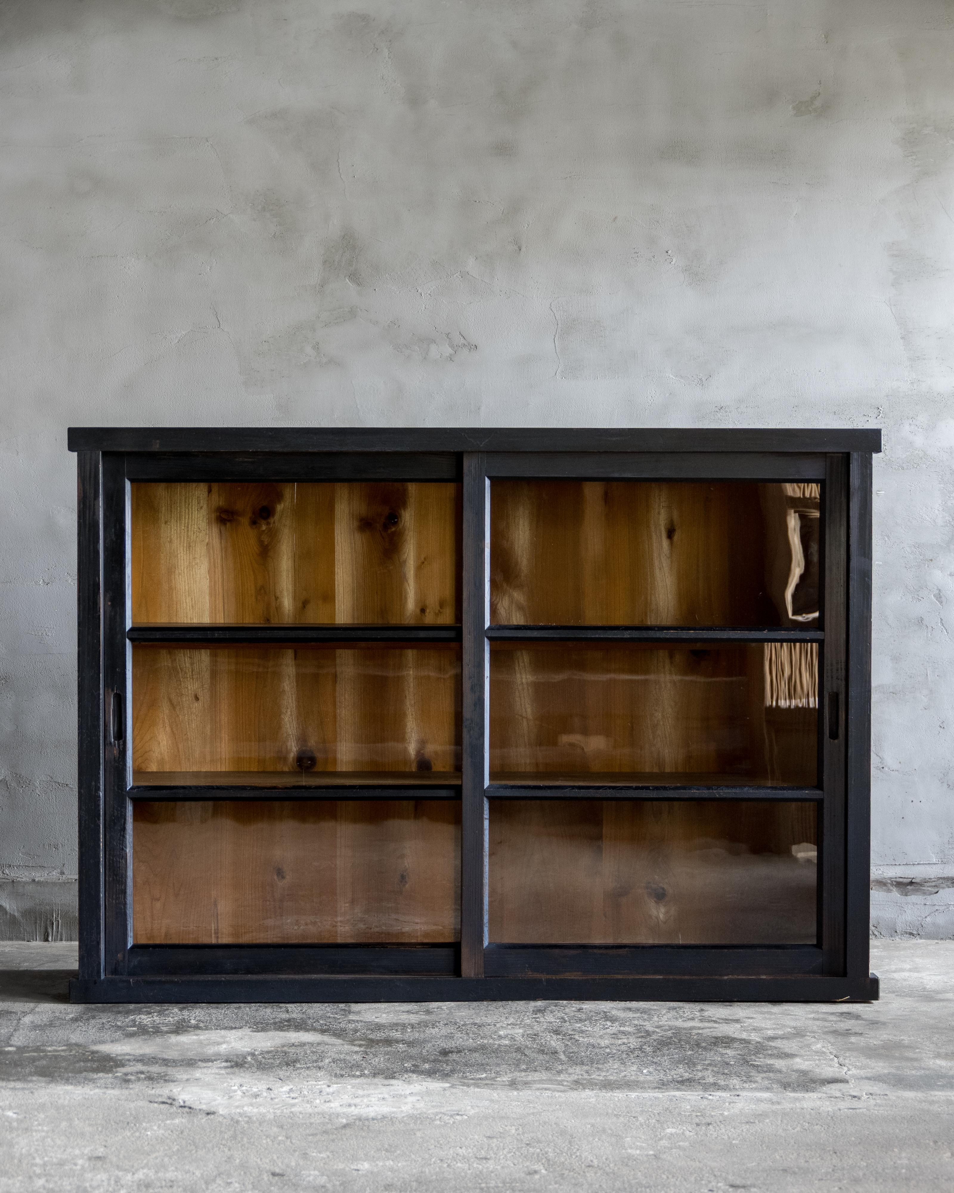 This vintage cabinet was crafted in the Early Showa Period (1926–).

Made from Japanese cedar with a black finish, the surface shows a gently lived-in patina that lends quiet wabi-sabi character without visual weight.

Behind the glass sliding doors