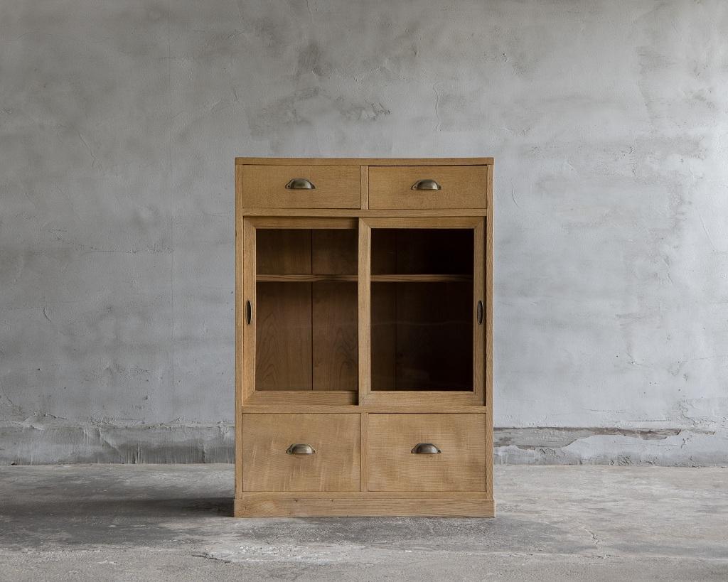 Este armario vintage japonés se fabricó a principios del Periodo Showa (1926-). 

Fabricado en madera Sen japonesa, tiene puertas de cristal e incluye cajones tanto en la parte superior como en la inferior, ofreciendo una excelente funcionalidad. La