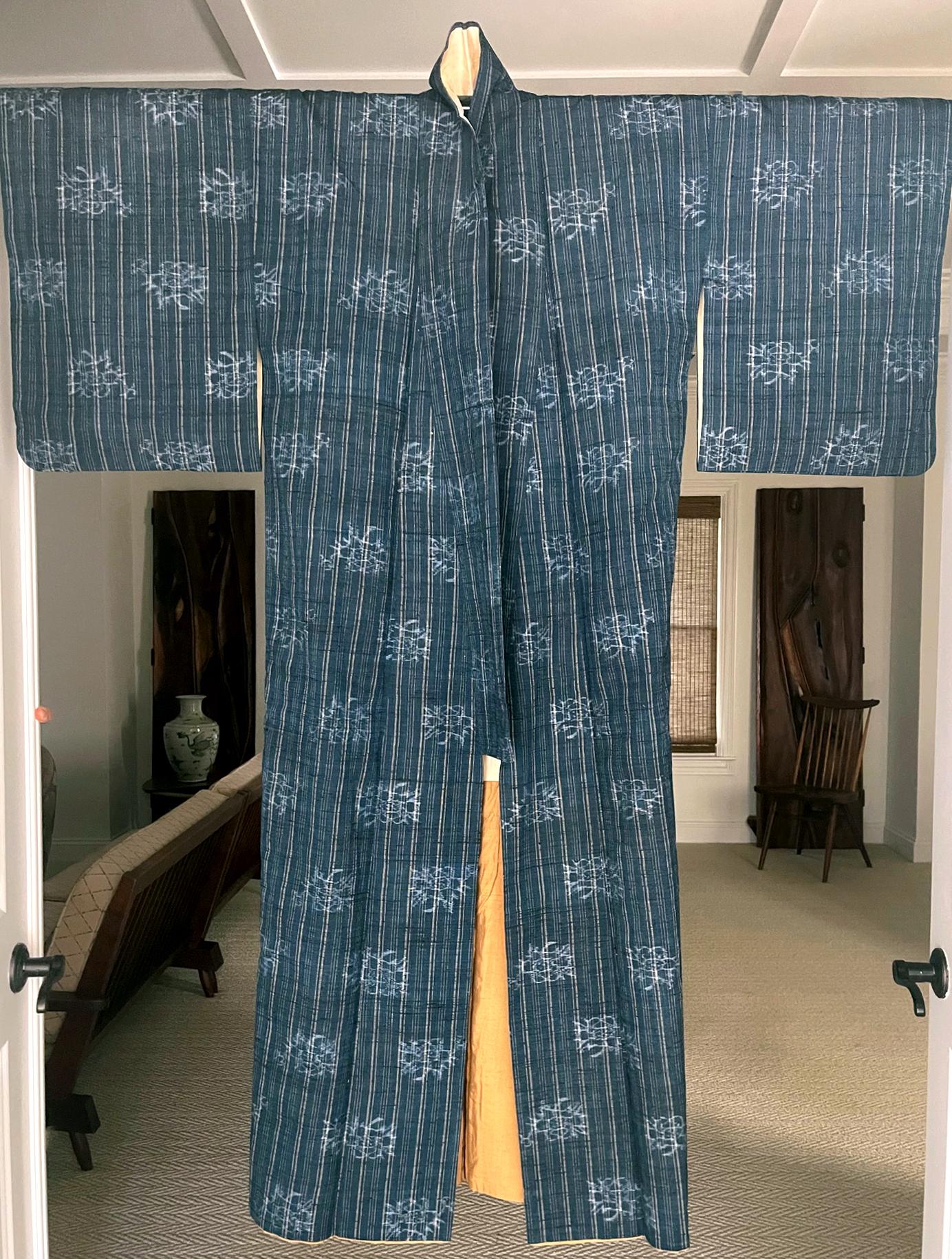 On offer is a Japanese summer kimono woven from indigo blue linen with ivory color fine stripes patterns with apparently darker weft additions. The elegant geometrical pattern is accentuated with stenciled white pattern of a floral design, likely
