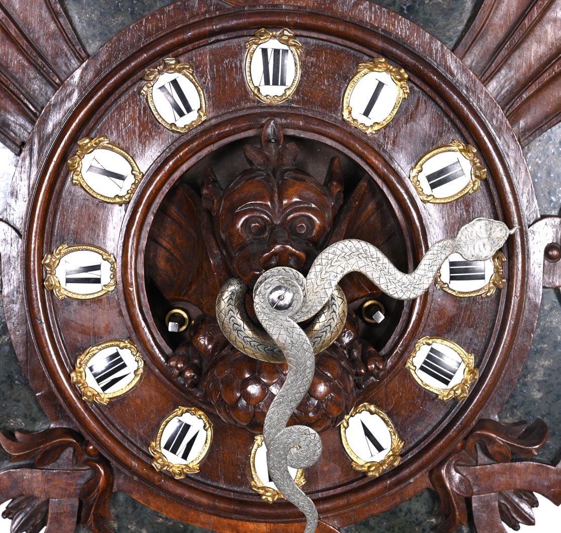 Eine auffällige japanische Wanduhr aus dunklem, geschnitztem Holz, verziert mit Intarsien aus tiefgrünem Hartstein. Die strahlende, fächerförmige Struktur ist mit fantastischen und mythologischen Figuren geschmückt.

In der Mitte hält ein
