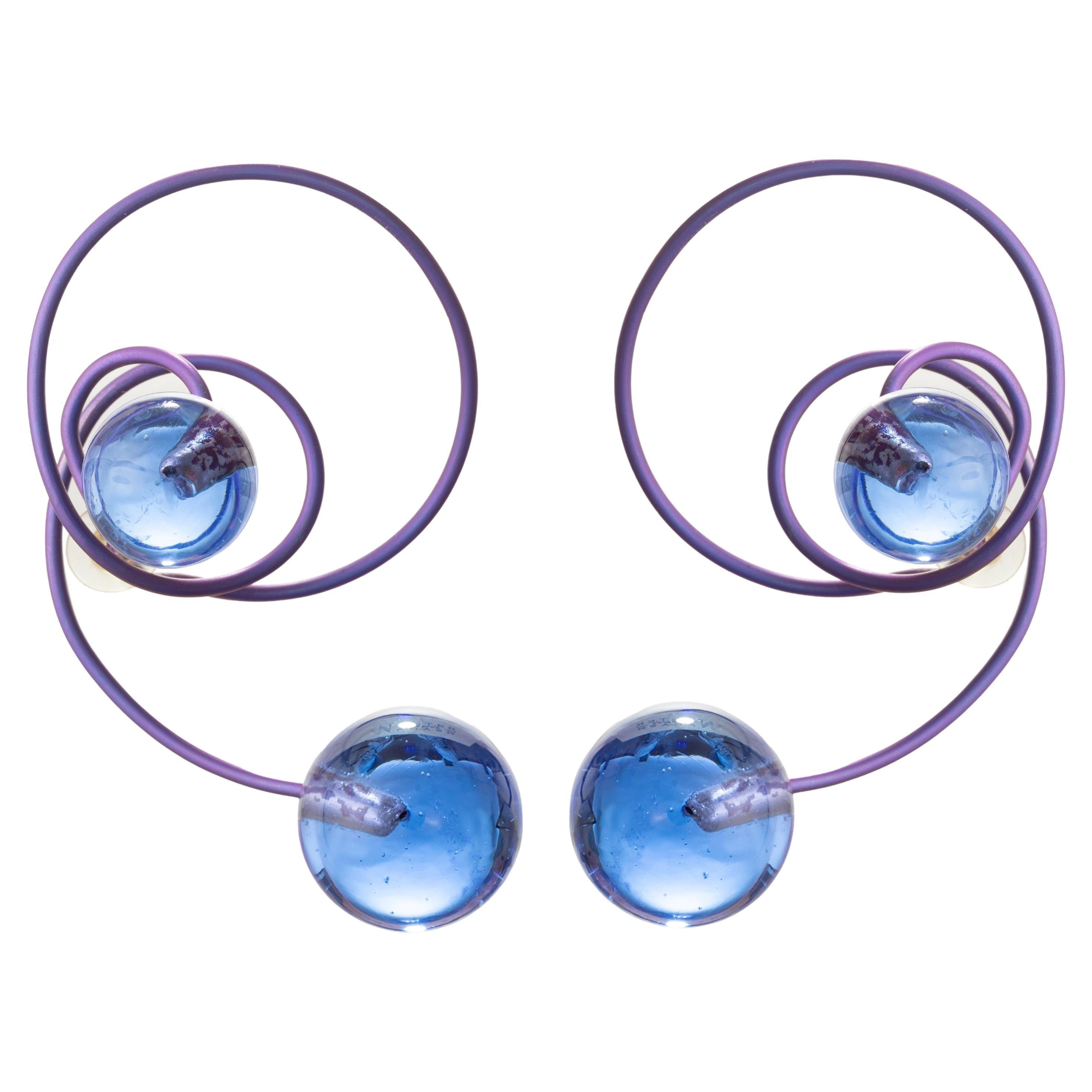 Playful titanium and Venetian glass earring creations by JAR Paris, featuring spirals of titanium in a shimmering purple shade with violet-blue glass bead end caps.  The earrings gently wrap and contour the ear, giving a visual symphony meant to