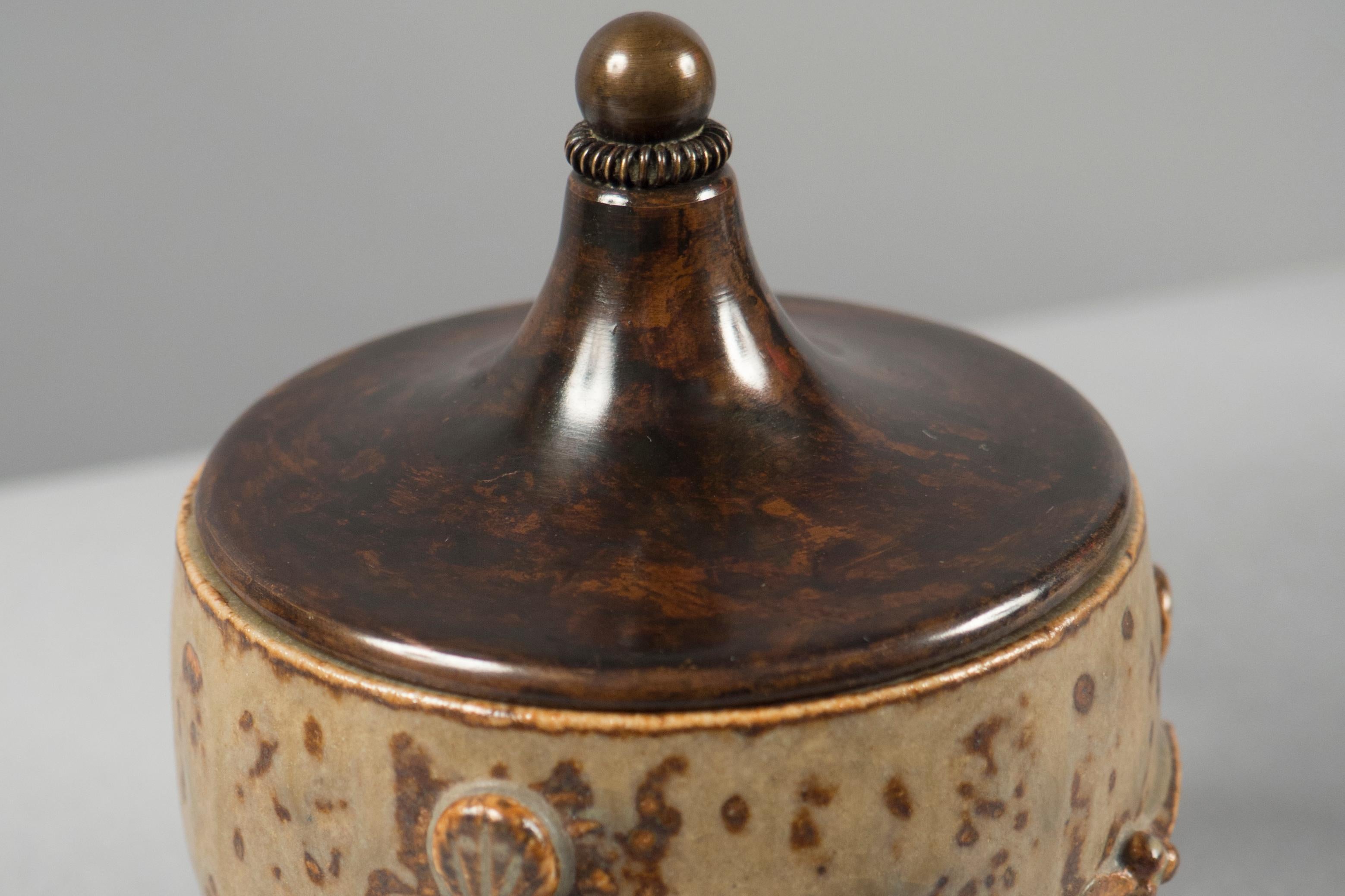 Brown glazed vessel with dark brown speckles with a raised floral motif throughout a patinated bronze lid tops it.