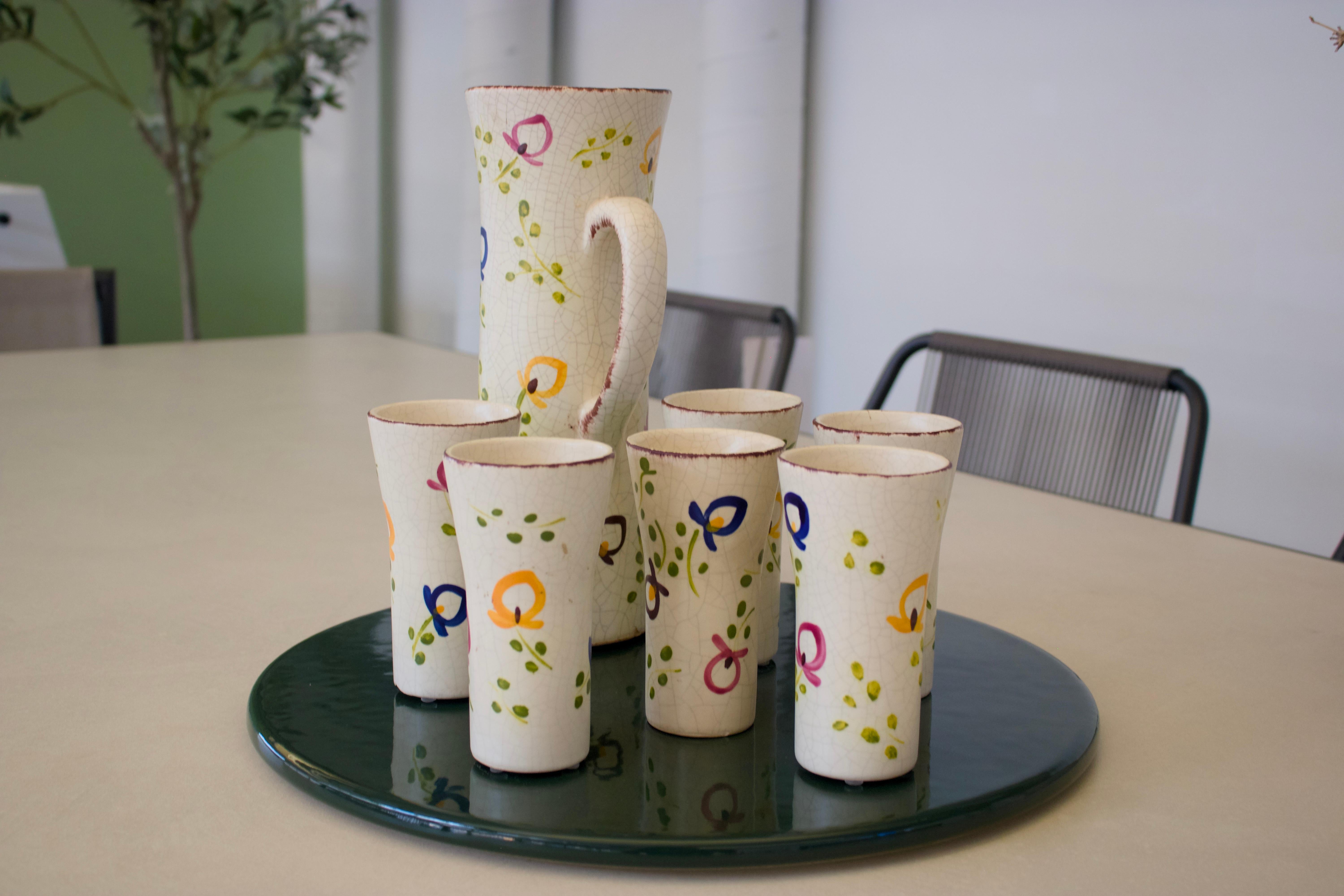 Hand-painted French ceramic carafe and set of six cups from the 1950s. These pieces capture the joie de vivre of mid-century with whimsical hand-painted motifs, charming circular patterns in vibrant blues, warm oranges, and deep burgundy all
