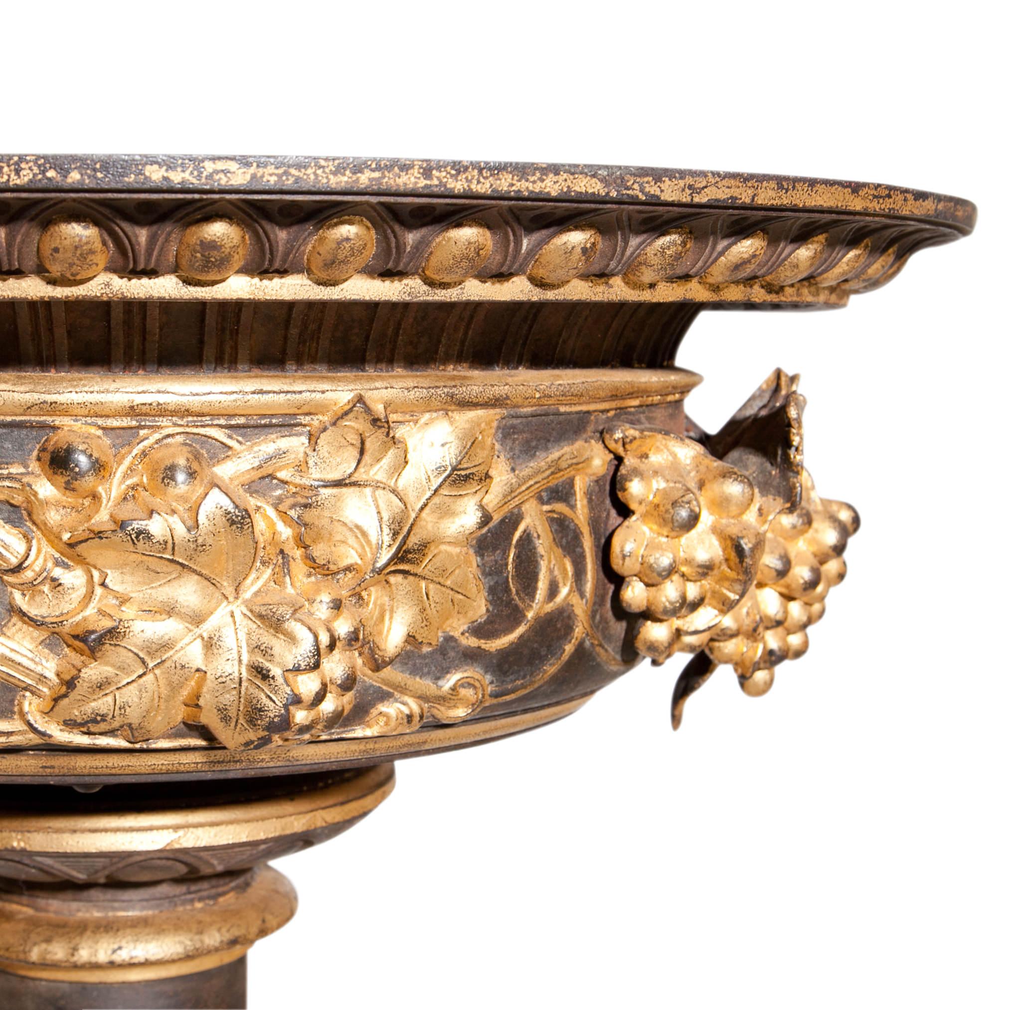 Large cast iron jardinière on a round stand with a square plinth, decorated with acanthus leaves. The basin is decorated with plastic grapes and vines in relief on the wall. Partly raised in gold.