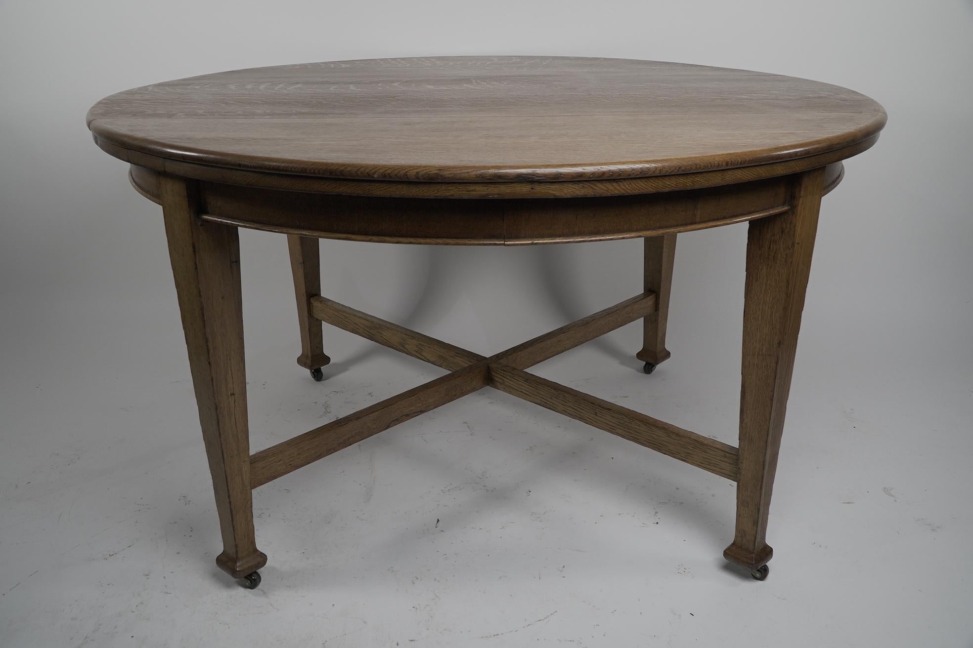 Jas Shoolbred London. An Arts and Crafts round oak dining table with square tapering legs united by a cross stretcher. Jas Shoolbred company disc attached underneath the table top.