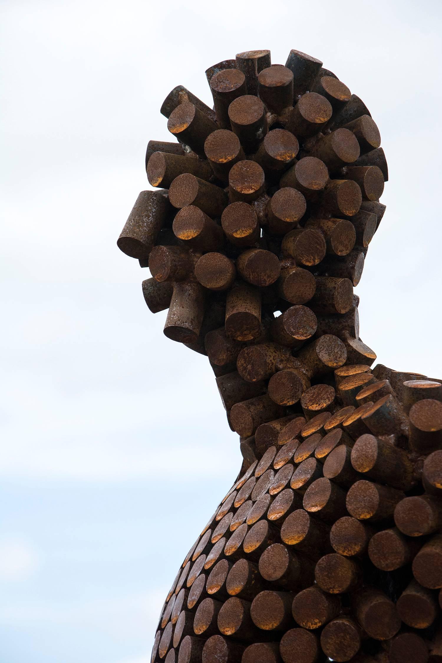 A seated figure reclines atop a steel cube in this outdoor sculpture by Mississippi artist Jason Kimes. Made of welded coin-sized discs of steel, the figure's shell is slightly transparent. The work that has taken on a weathered, rusted patina