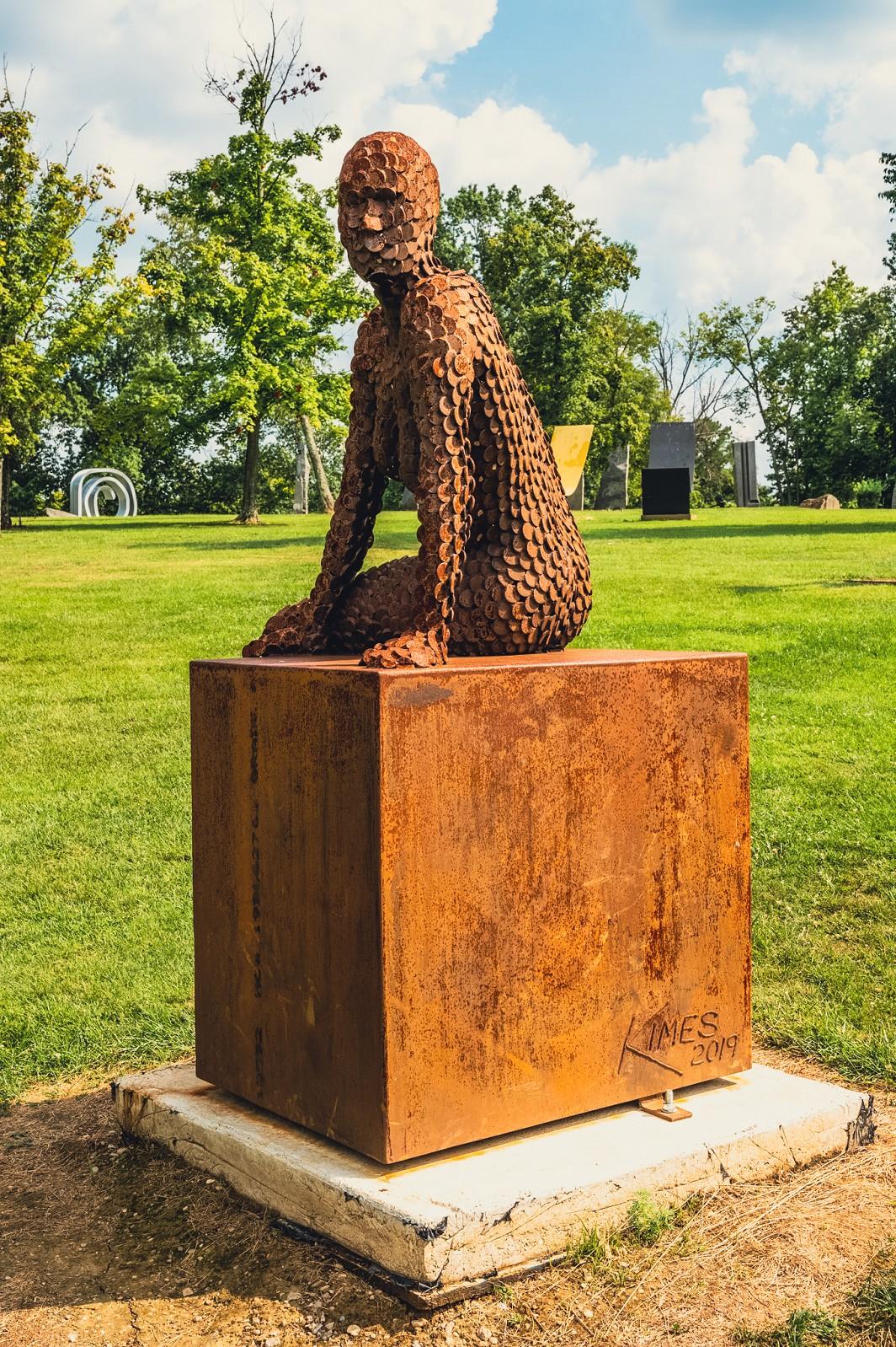 A seated female figure strikes a casual pose, body half turned in this imposing sculpture by American artist, Jason Kimes. This outdoor sculpture is made from coin-sized discs of corten steel which naturally weathers over time acquiring a beautiful