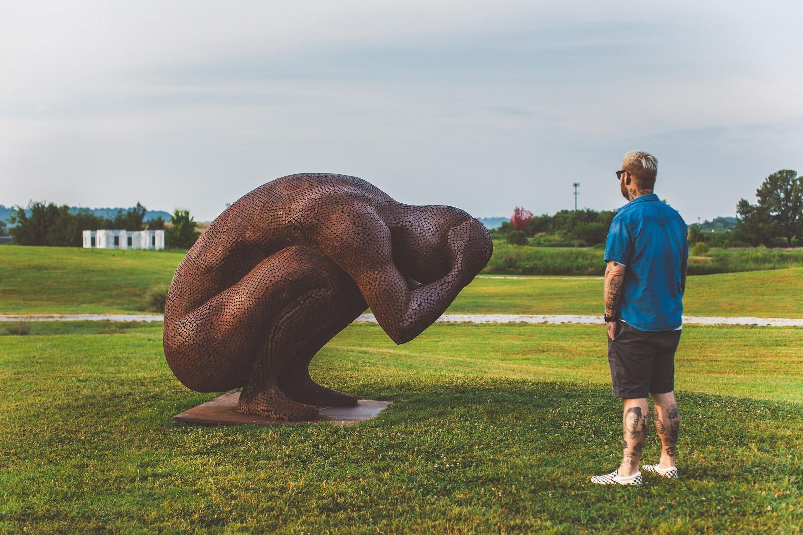 A figure squats, head held in his hands in this dramatic and monumental steel sculpture by Mississippi artist Jason Kimes. This outdoor sculpture is made from coin-sized discs of corten steel which naturally weathers over time acquiring a beautiful