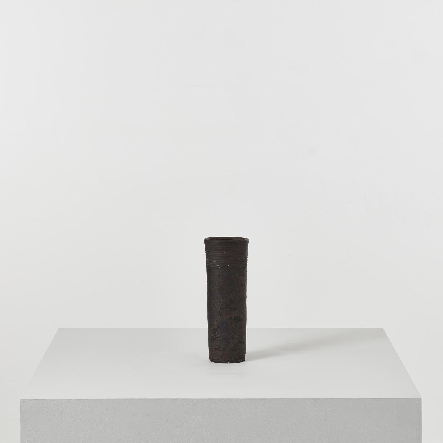 De forme cylindrique, réduit à l'état d'ornement, ce vase Jason Wason se délecte de la matérialité de l'argile. Avec une série de bandes horizontales nervurées tactiles sur le col, la surface patinée et texturée du récipient a un aspect élémentaire.